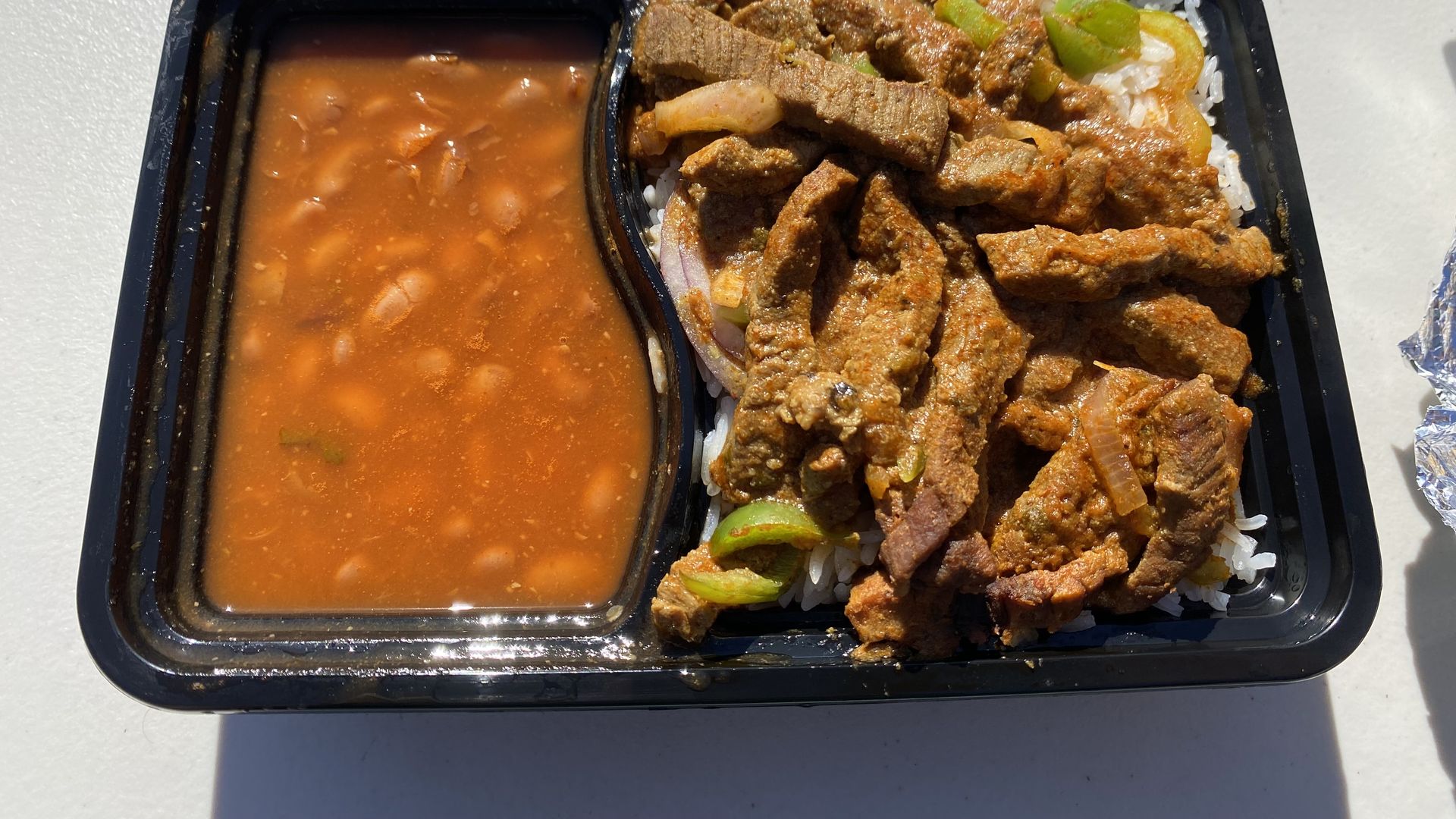 A compartmentalized plastic container with beef strips over rice on the right side and pinto beans on the left.
