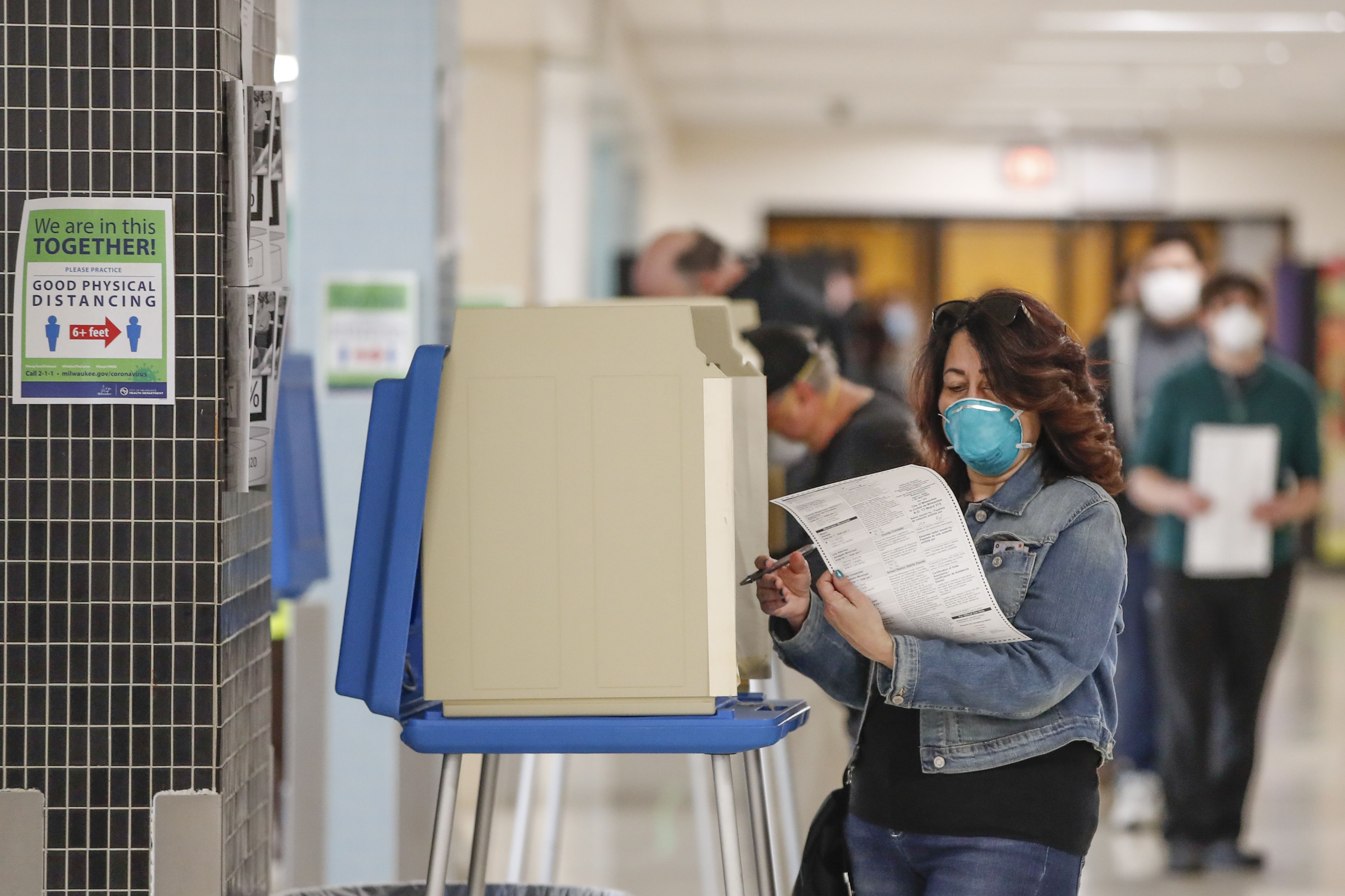 In this image, a woman wears a face mask and fills out a sheet of paper