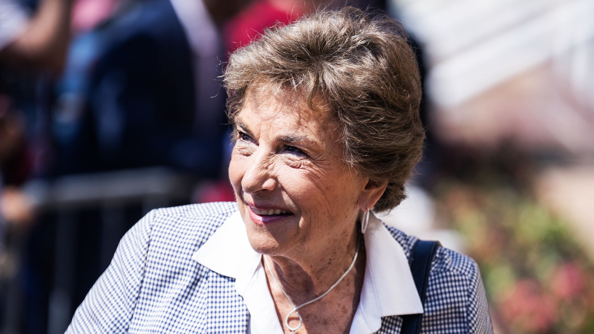 Smiling Jan Schakowsky with short brown hair wearing a checkered blazer, white shirt, pearl necklace, and a pin on her lapel, outdoors with blurred background.