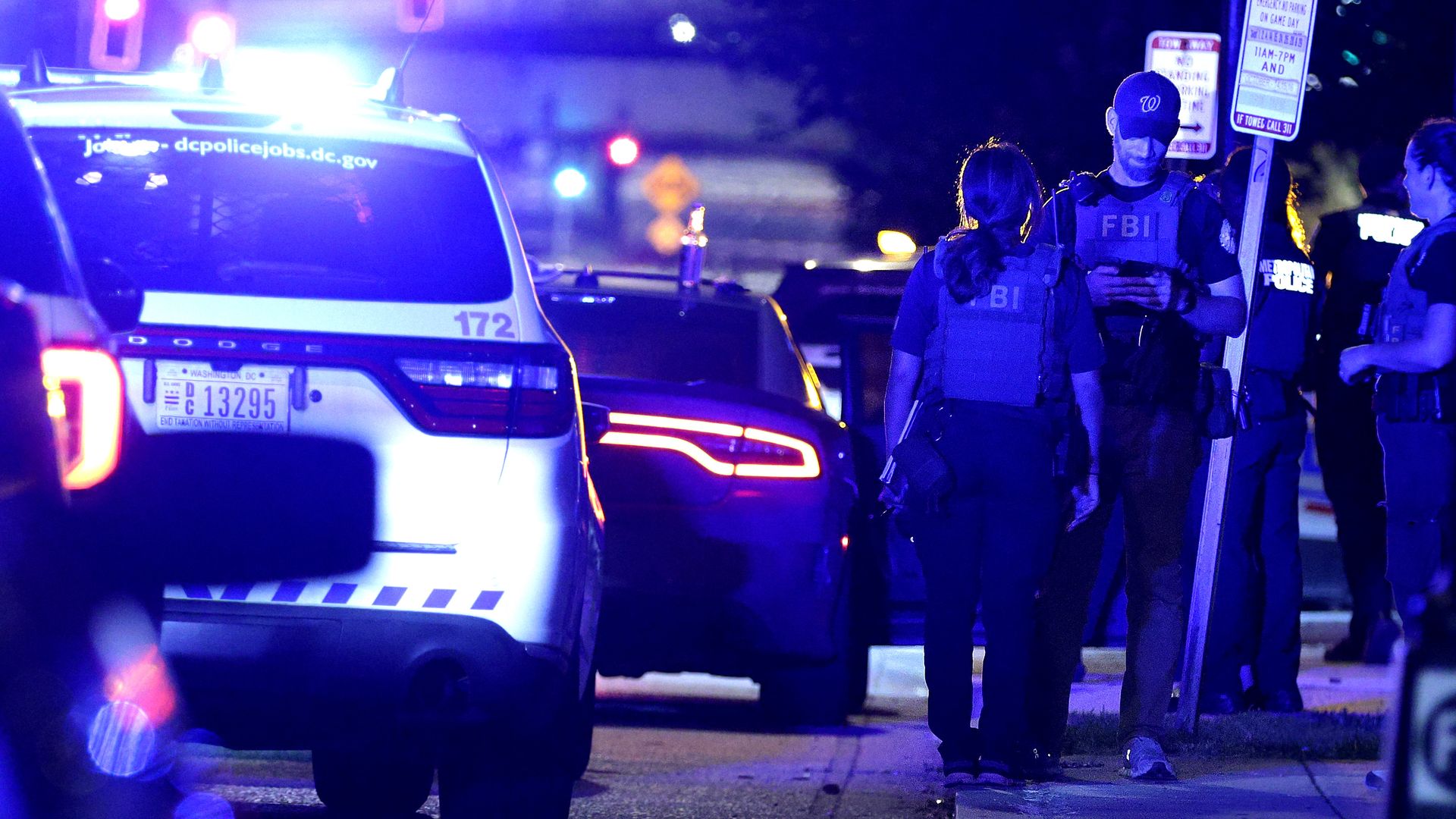 FBI agents and police officers at a nighttime scene with a parked police SUV, another vehicle, and bright blue emergency lights illuminating the street.