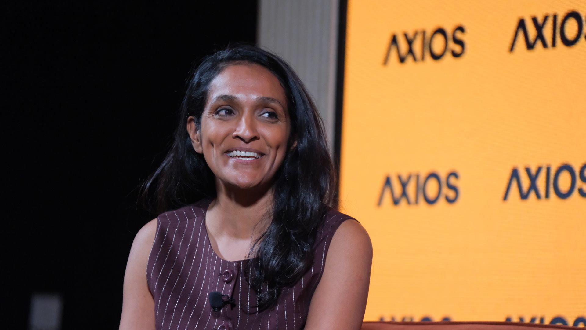 Smiling mayoral candidate Nithya Raman in a maroon pinstripe dress sits on stage with a large orange Axios backdrop to the right; a small microphone is clipped to her chest.