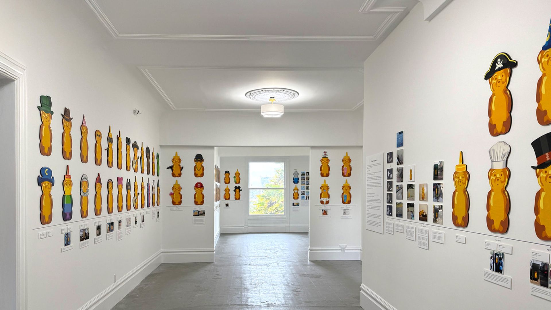 Art gallery room with white walls displaying multiple orange peanut-shaped figures wearing various colorful hats, costumes, and accessories, accompanied by small placards and photos.