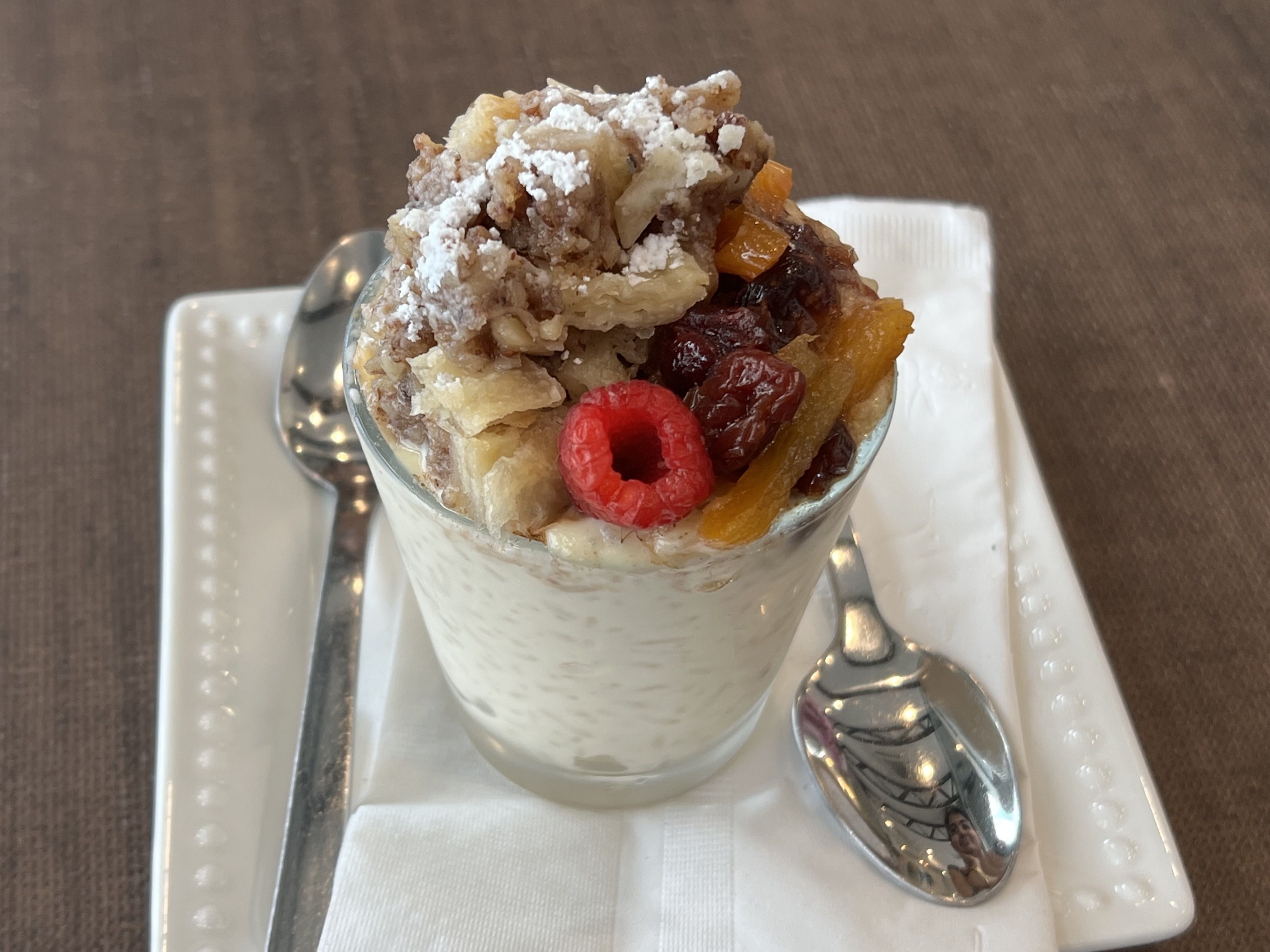 Glass of creamy rice pudding dessert topped with powdered sugar, mixed nuts, dried fruits, and one fresh red raspberry, placed on a white plate with two silver spoons and a white napkin on a brown table.