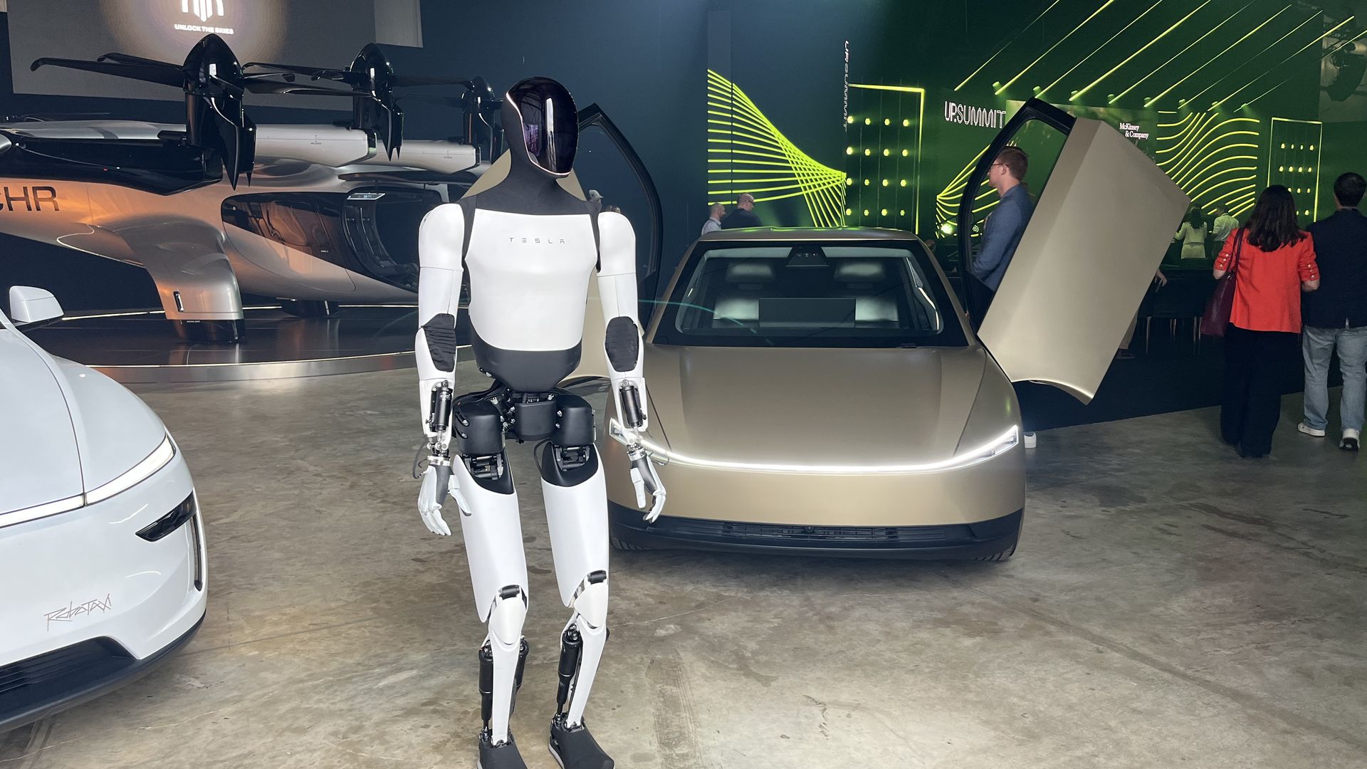 White and black Tesla humanoid robot standing indoors near a gold Tesla robotaxi with gull-wing door open. Behind them is a futuristic electric air taxi from Archer.