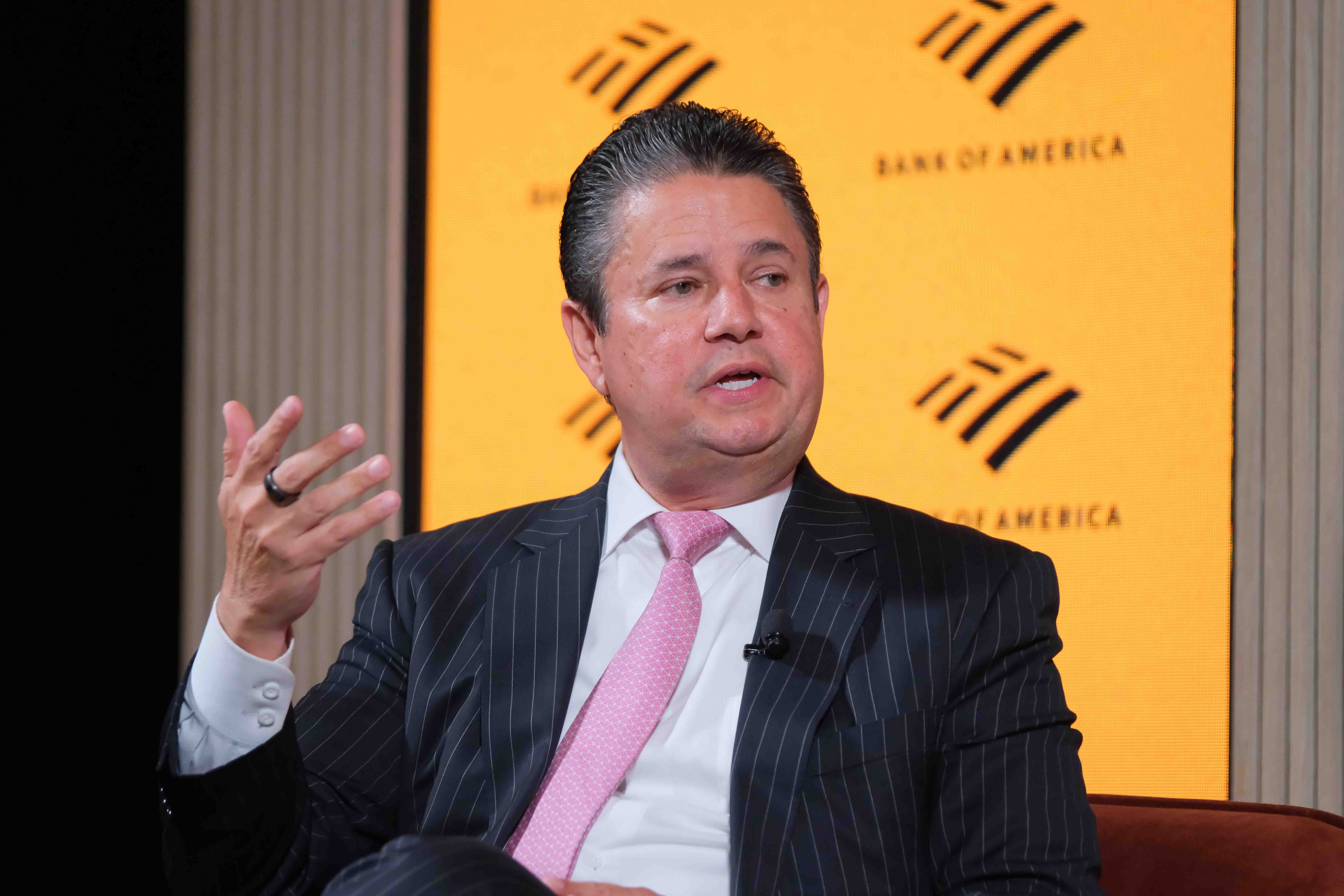A man in a dark pinstripe suit and pink tie speaks at a panel, gesturing with his hand; a bright yellow backdrop with diagonal black lines is behind him.