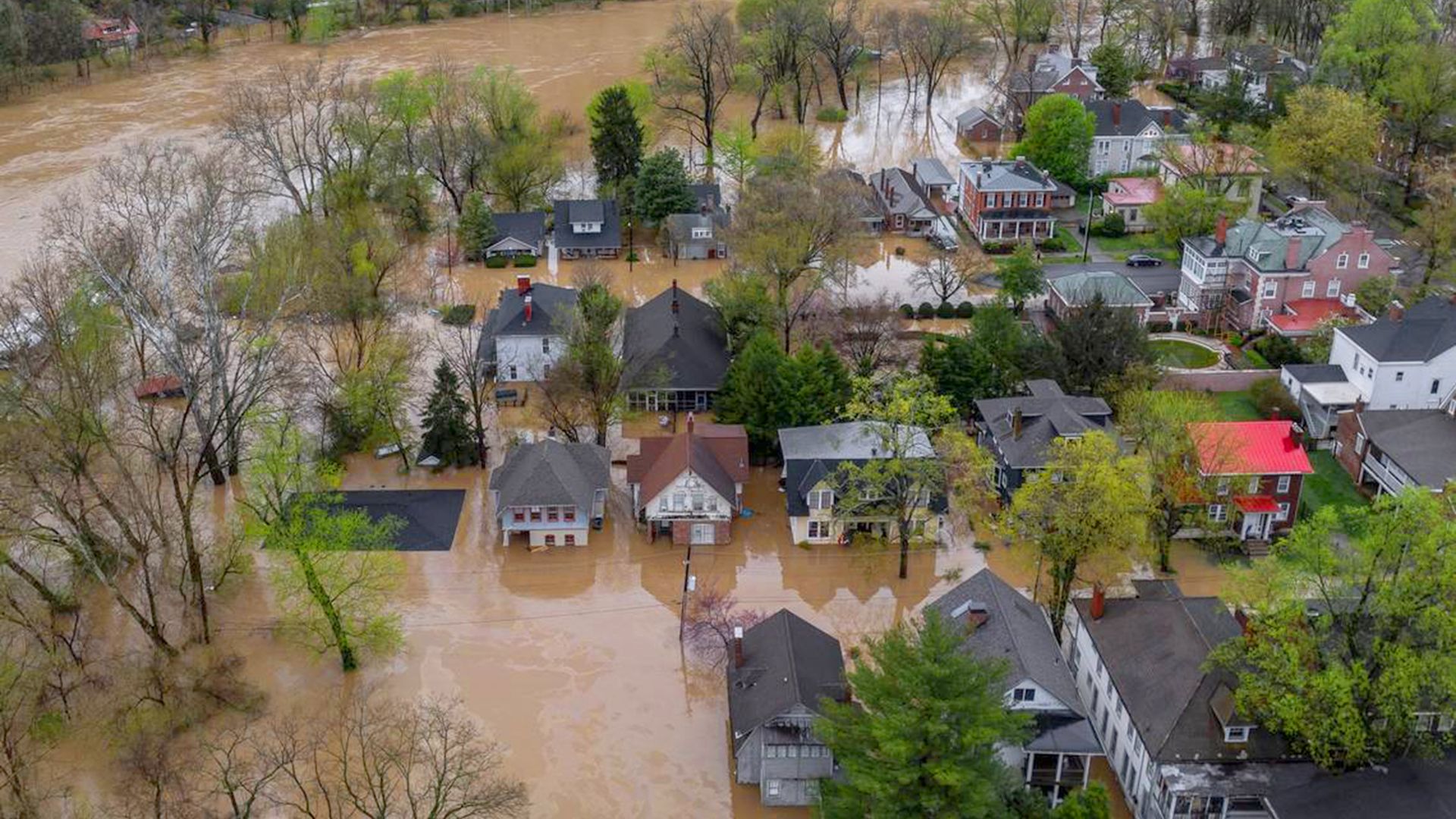 Aerial view of a flooded neighborhood, with muddy water surrounding homes, streets, and trees near an overflowing river.