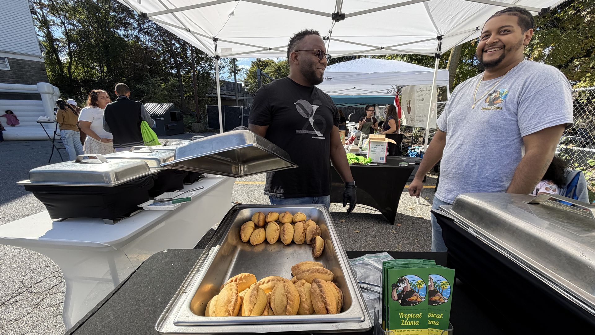 Johan Martinez, left, lifts up a tray to show the Bolivian mini-empanadas from Tropical Llama being sold under the white canopy in a Roslindale parking lot at a Hispanic Heritage Month cookout.