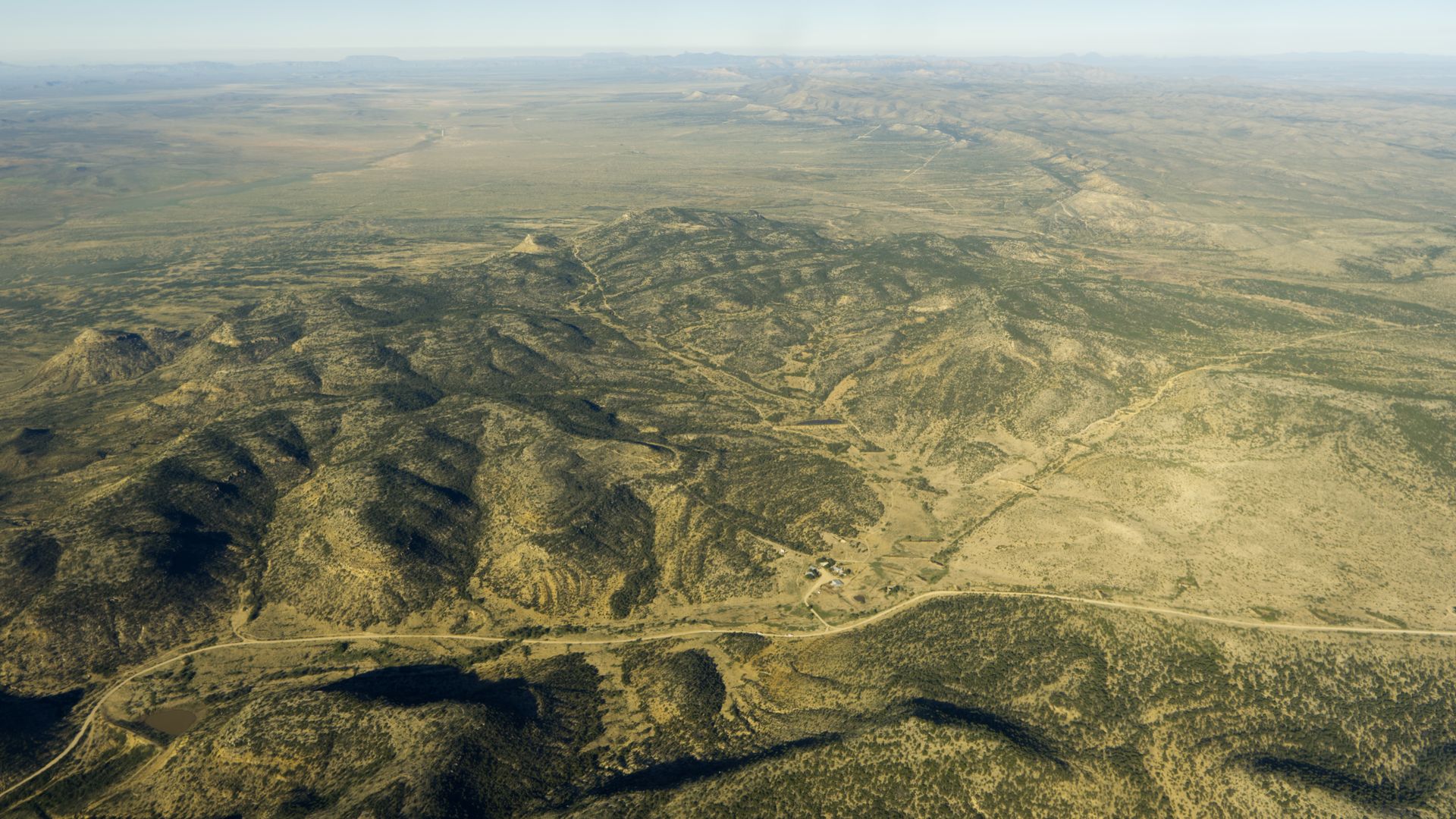 A bird's eye view of a remote part of Texas. The hills are green and brown. The sky is hazy.