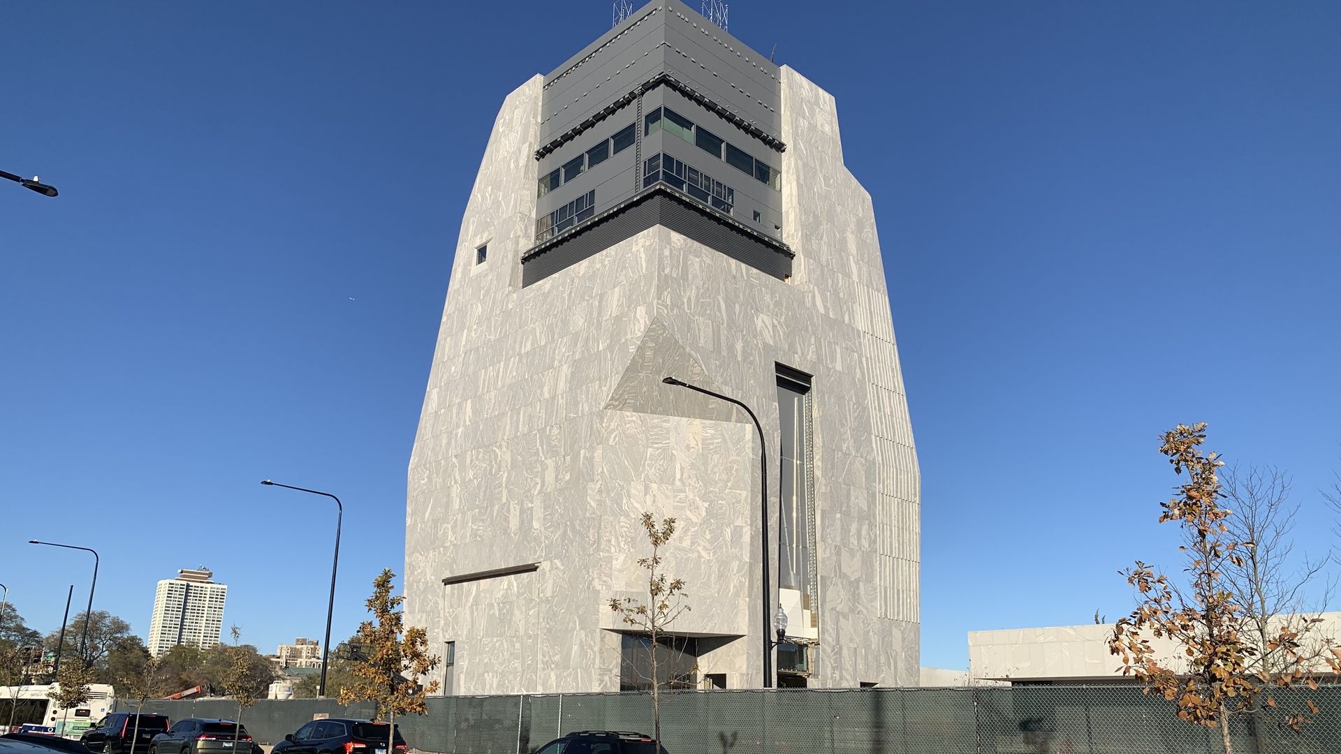 Tall, angular building with light gray marble facade and black top section under clear blue sky, surrounded by cars, street lamps, young trees with autumn leaves, and a chain-link fence.