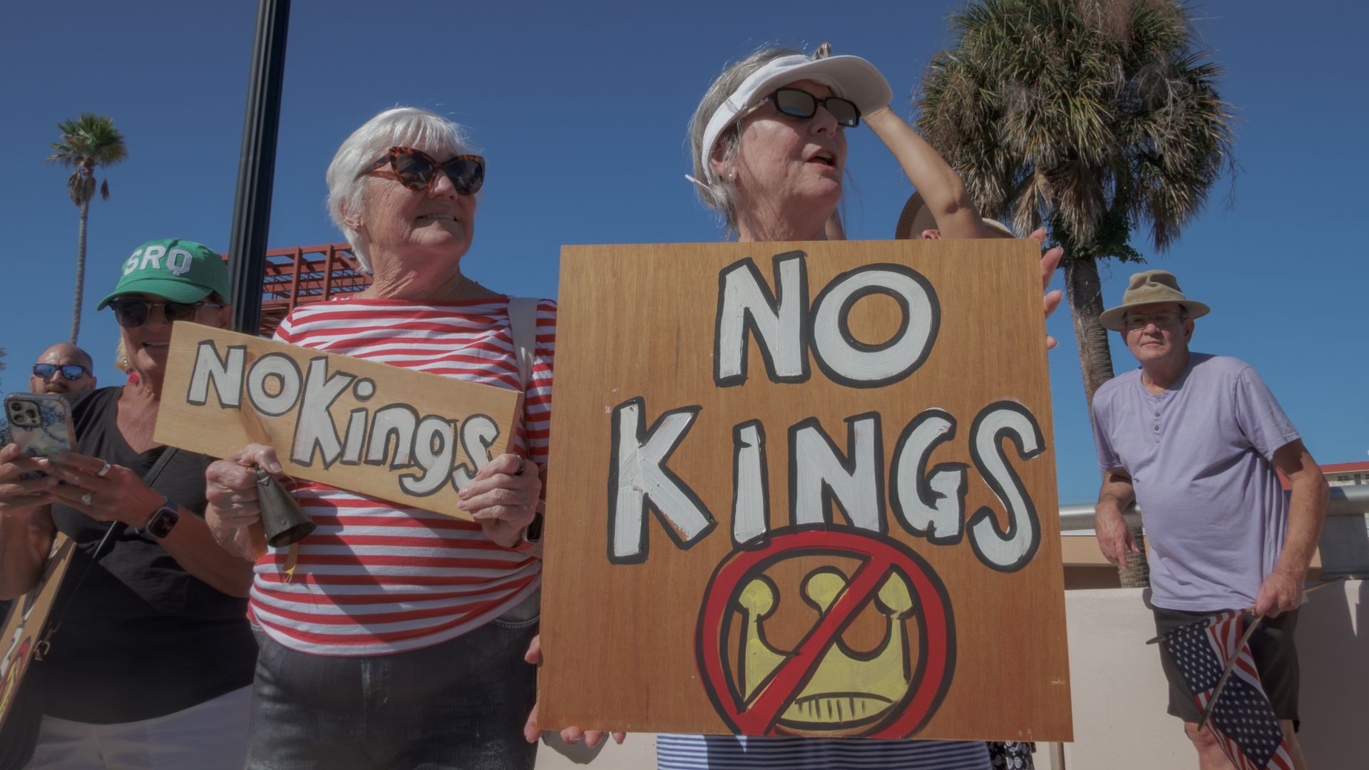 Group of protesters outdoors on a sunny day; two elderly women hold signs reading "NO KINGS" (one large wooden board with a crossed-out symbol), while a man in a hat and others watch.