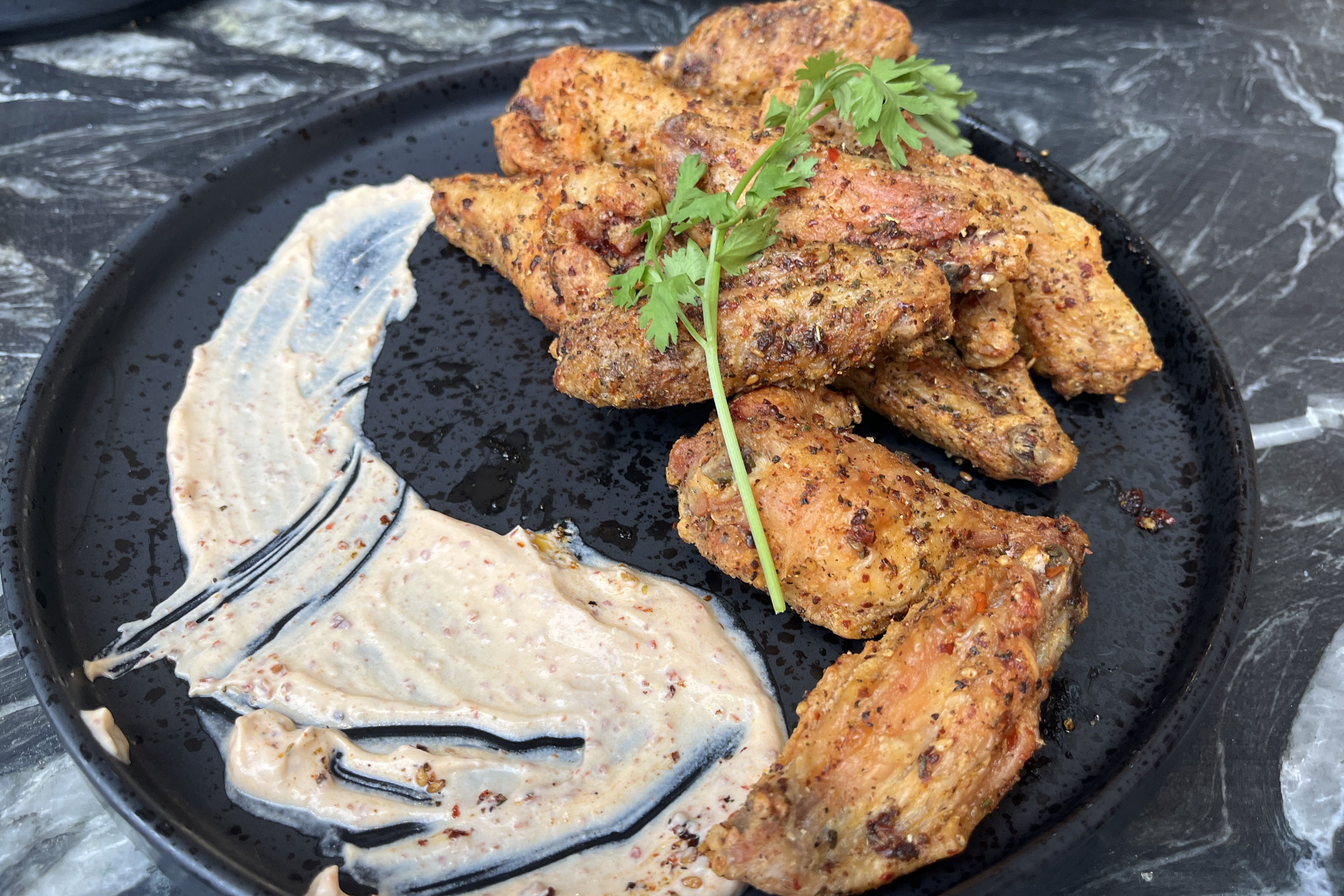 Crispy, seasoned chicken wings stacked on a black plate, topped with a parsley sprig, with a smear of creamy sauce on the side against a dark marble background.