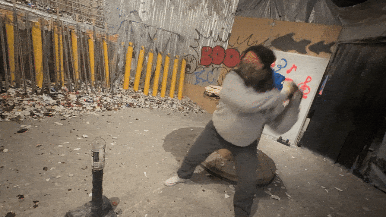 Person wearing gray clothing and a black hat swinging a bat inside a room with graffiti, yellow poles, and broken glass scattered on the floor, likely a smash room.