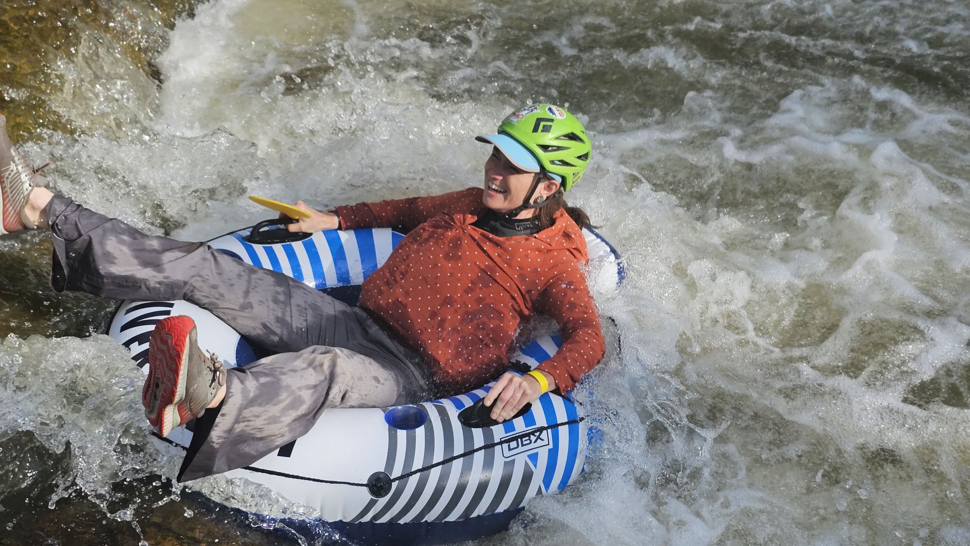 Smiling person wearing a bright lime-green helmet with a blue visor sits in a blue-and-white striped inflatable raft, holding a yellow paddle through rough whitewater.