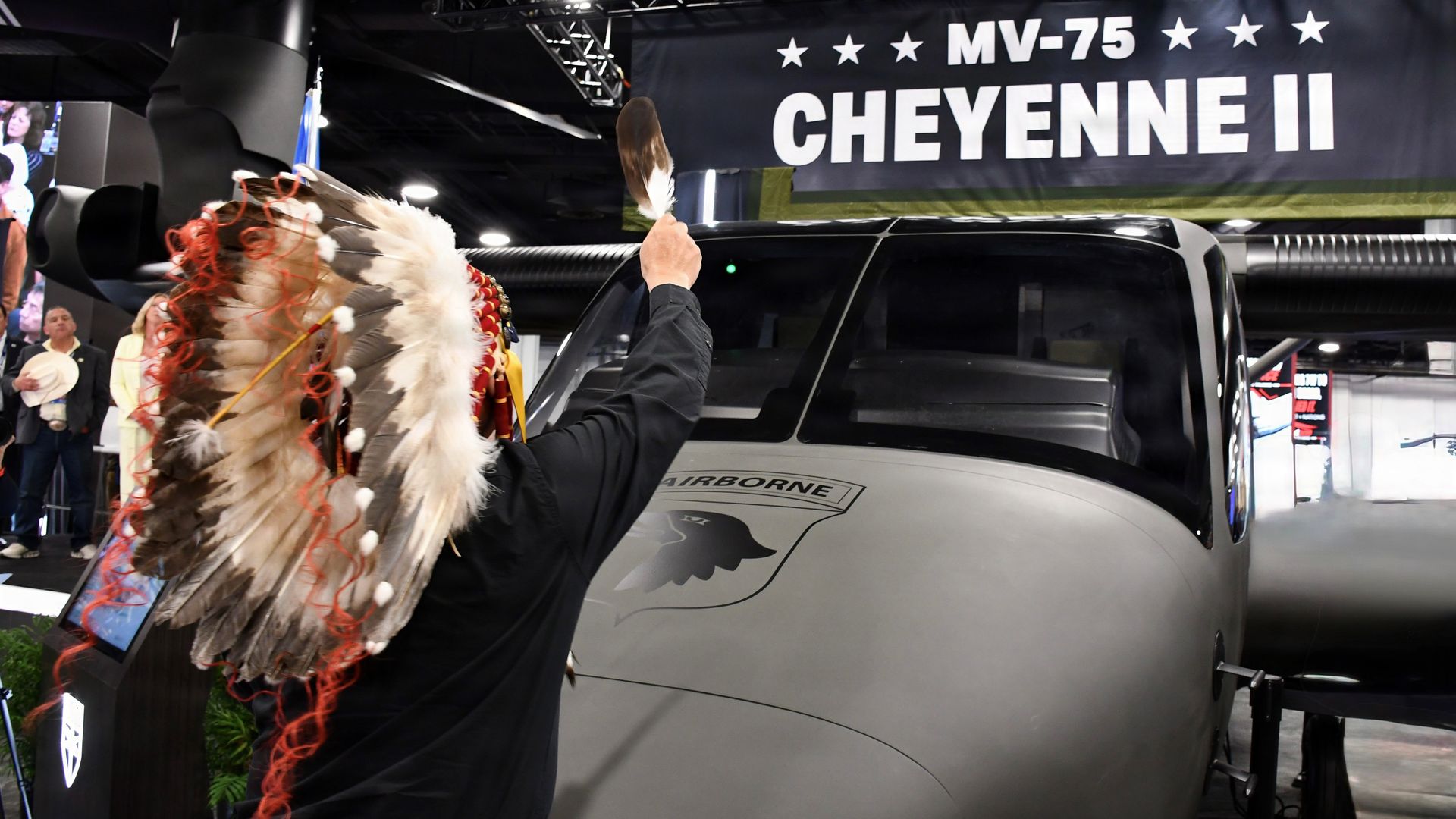 A defense conference scene, with a gray aircraft and a person in black raising a feather toward it. A banner overhead reads "MV-75 CHEYENNE II."