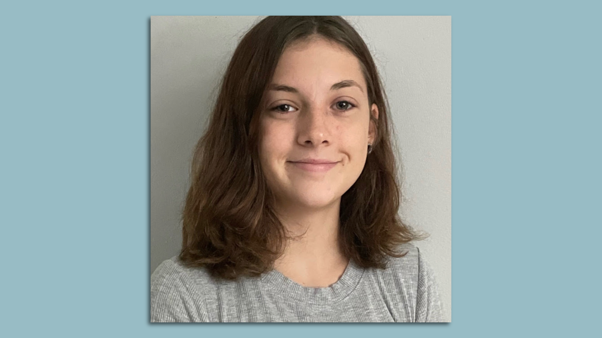 A teenage student with shoulder-length brown hair and wearing a gray shirt smiles at the camera on a blue background.