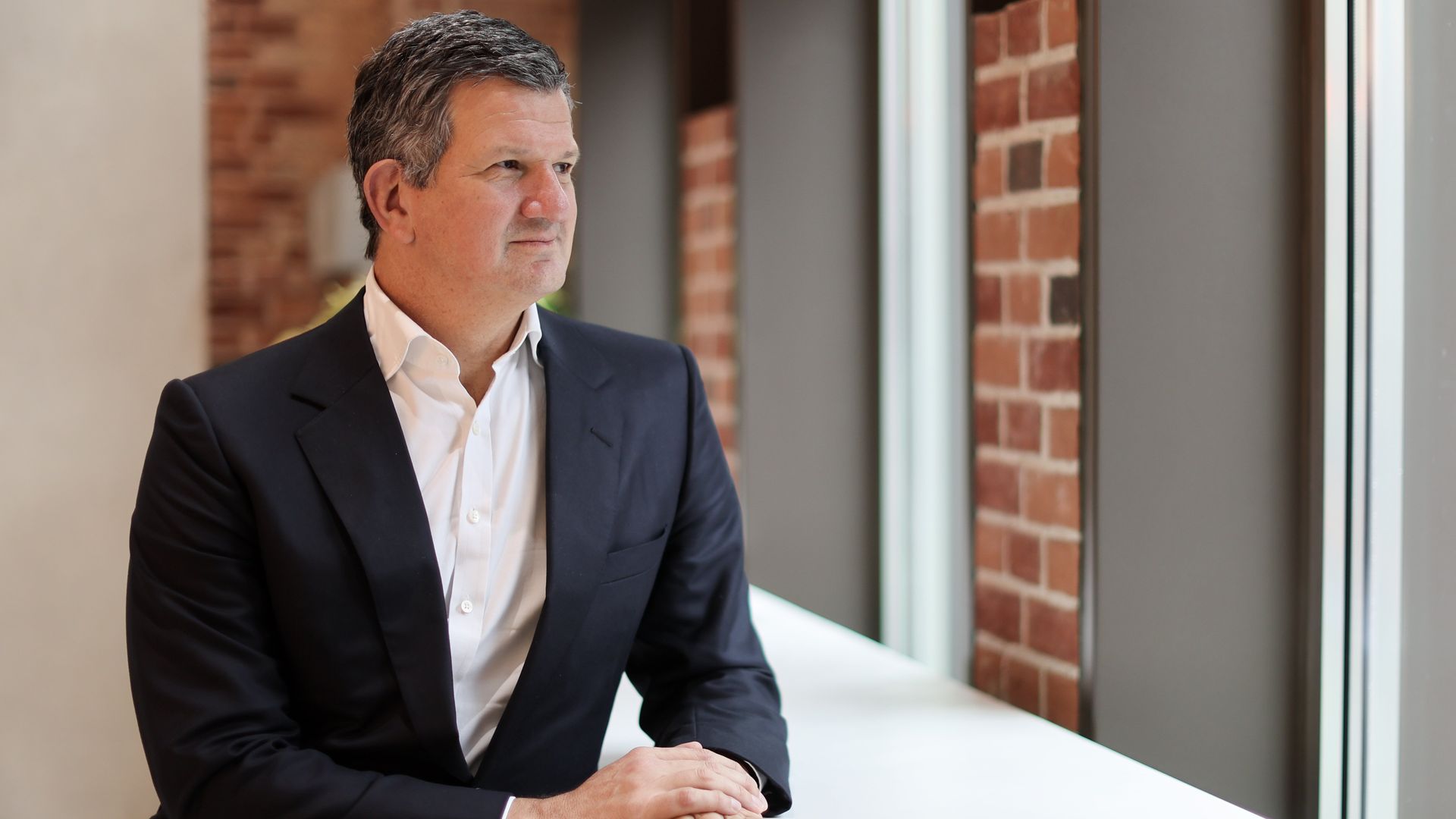 Steve Hasker, CEO of Thomson Reuters, in a dark suit and white shirt sitting at a white table, looking out a window with brick walls in the background.