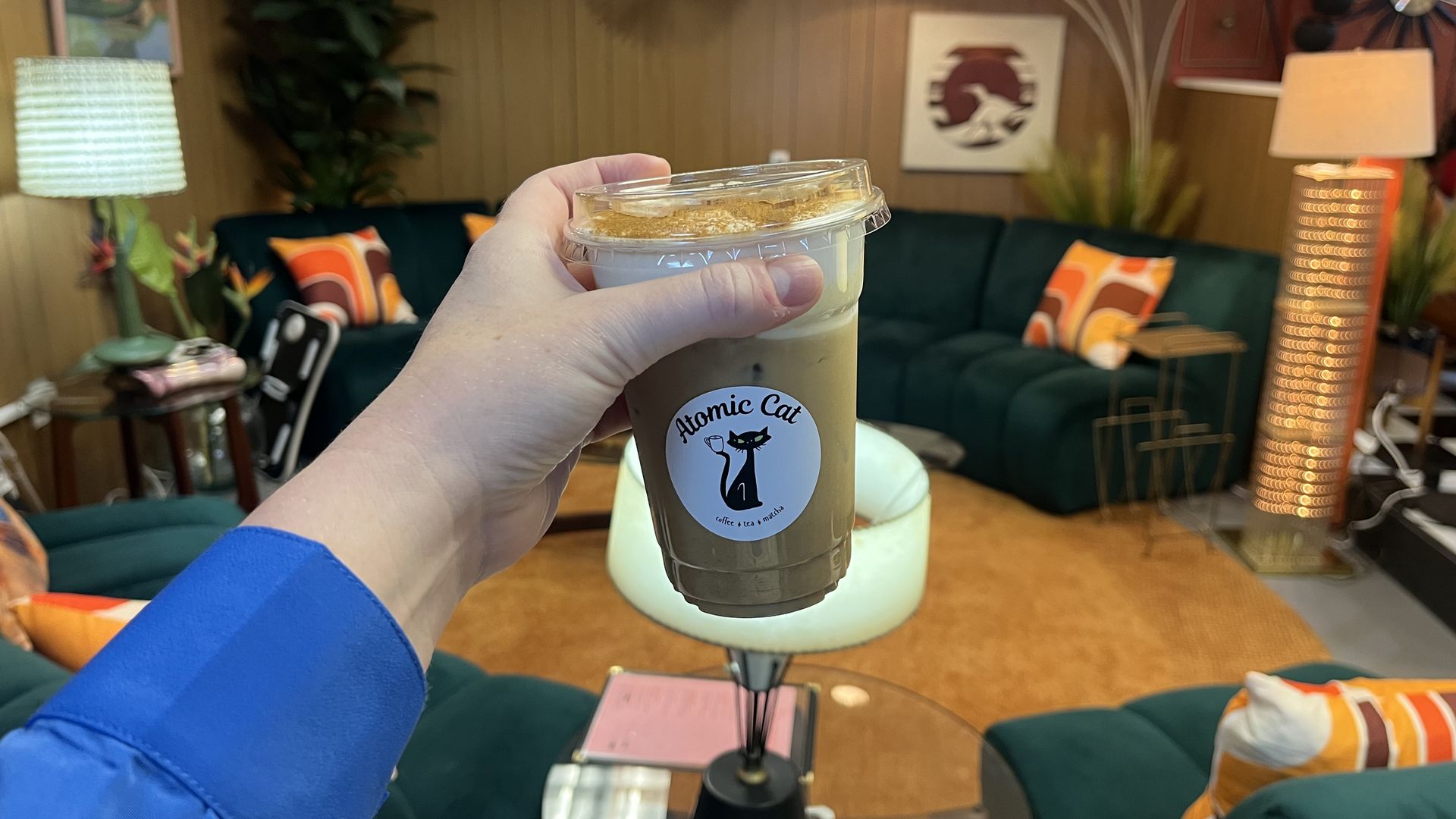Close-up of a hand holding an iced coffee cup labeled "Atomic Cat" in a retro living room with wood panel walls, a green sofa, colorful cushions, and mid-century lamps.