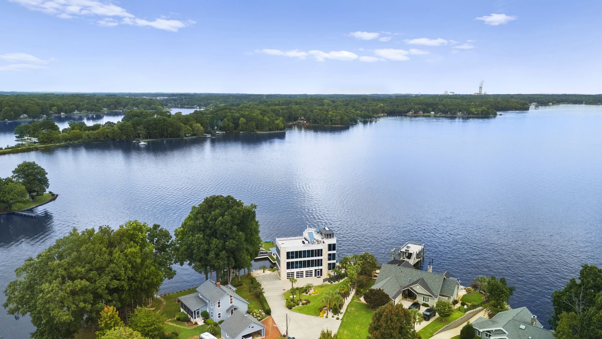 Aerial view of a lakefront neighborhood with multiple houses surrounded by trees and lawns along calm blue water under a partly cloudy sky.