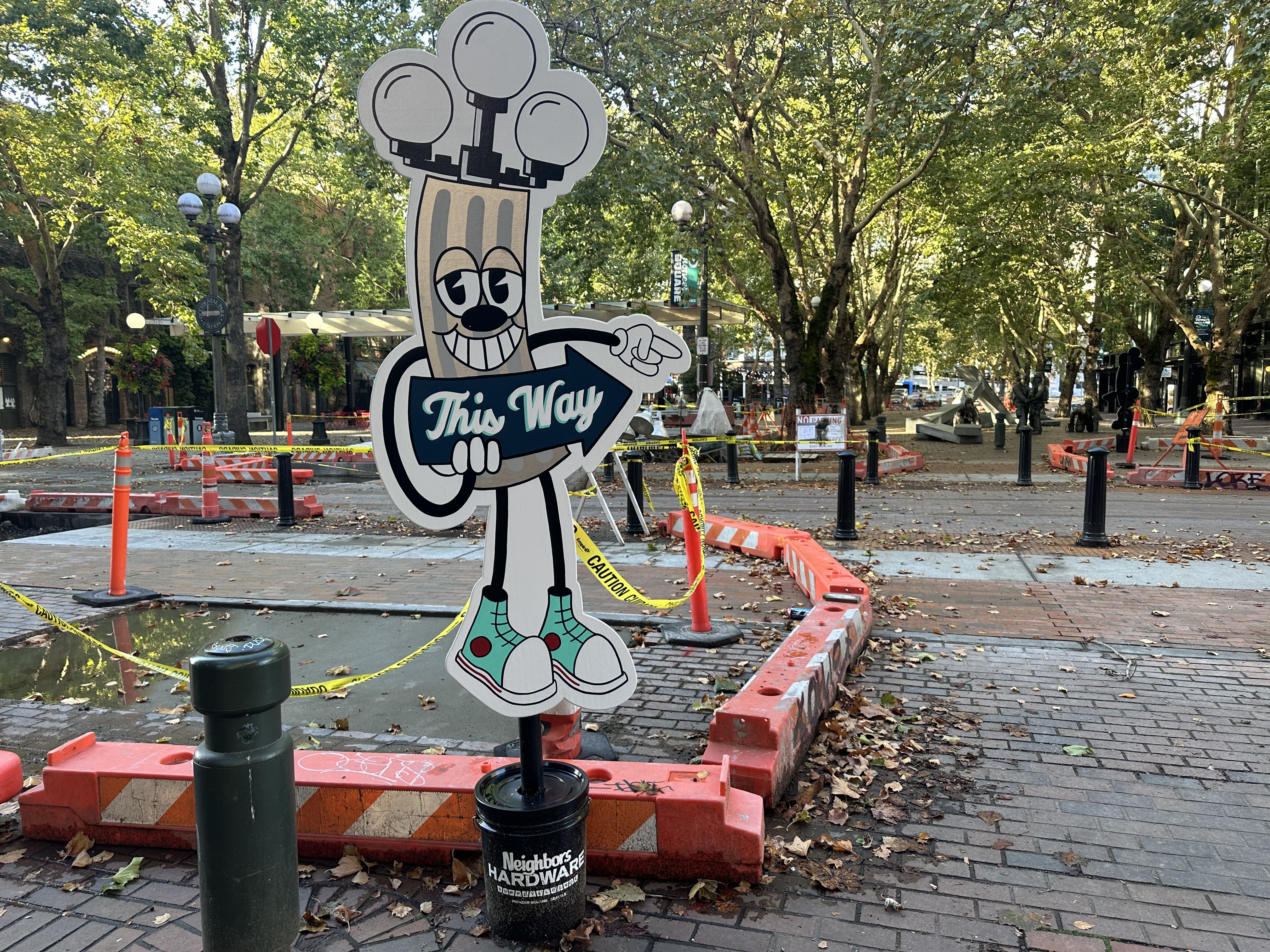 A cartoon cutout sign that appears to be a lamp post with legs, arms and shoes holds a sign that says "This way," pointing to an opening between construction areas,