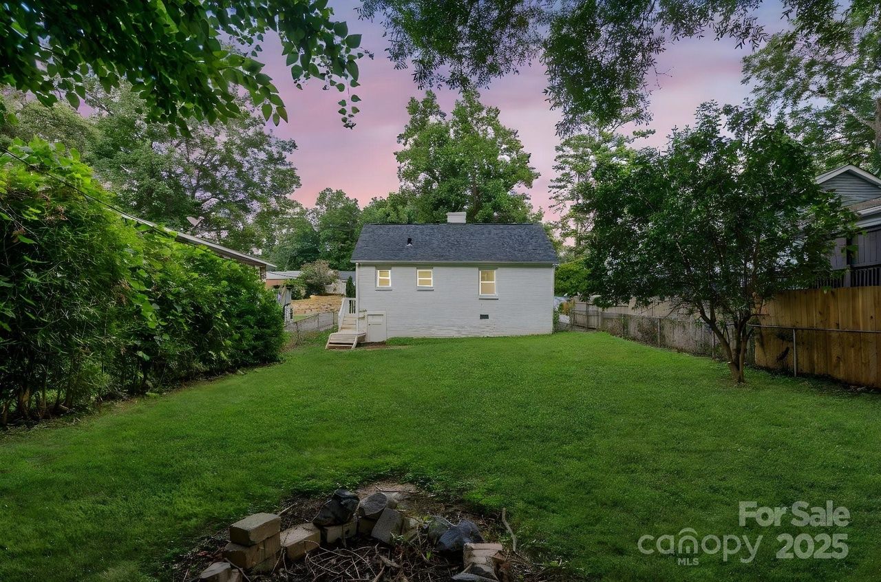 View of a small white house with a dark roof and lit windows at dusk, surrounded by a green lawn, trees, and a firepit made of stones in the foreground. Text in corner reads "For Sale canopy MLS 2025".