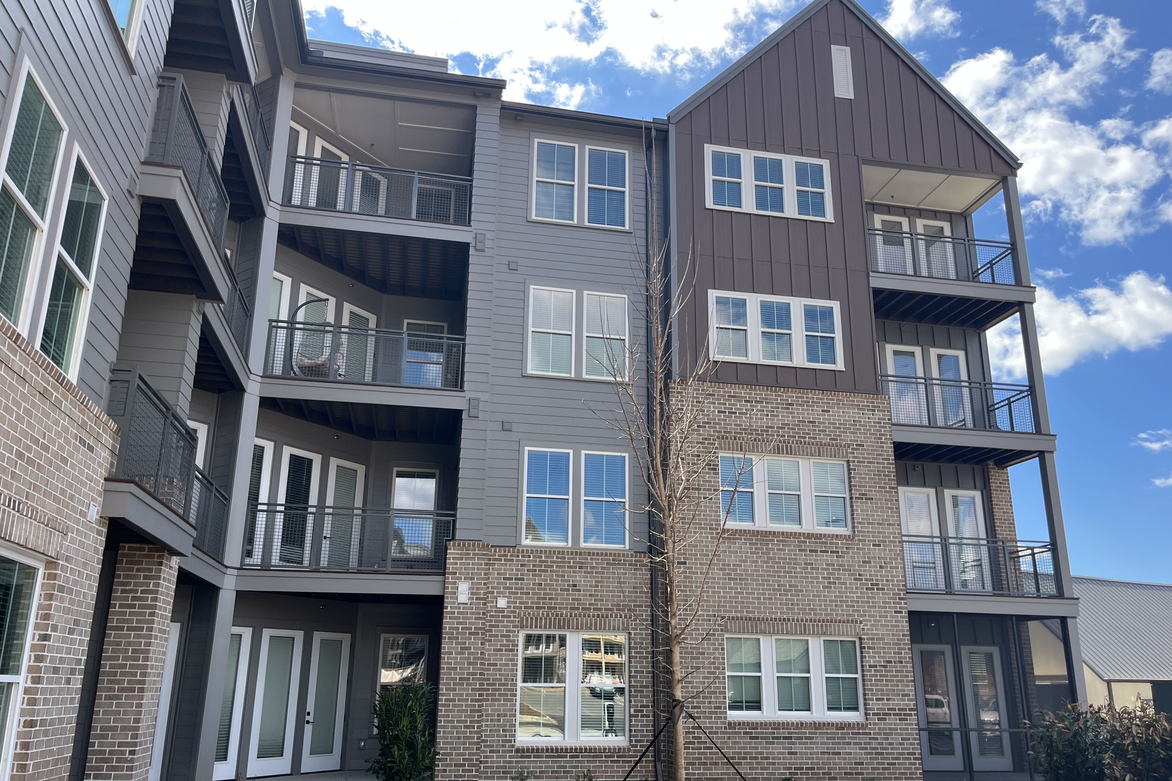Multi-story apartment building with gray siding and brick lower levels, several balconies with metal railings, large white-framed windows, a leafless tree in front, blue sky with clouds.