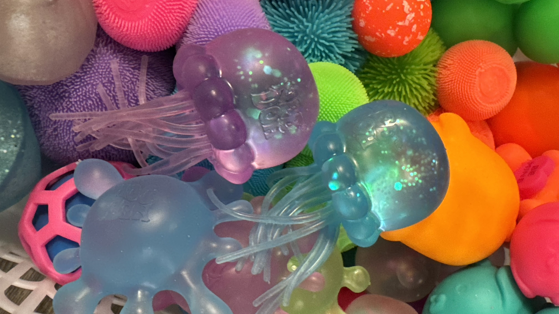 Bright collection of translucent jellyfish-shaped squishy toys in blue, purple, pink, and teal, with tentacles and rounded bodies, set on a white lattice surface among colorful textured balls.