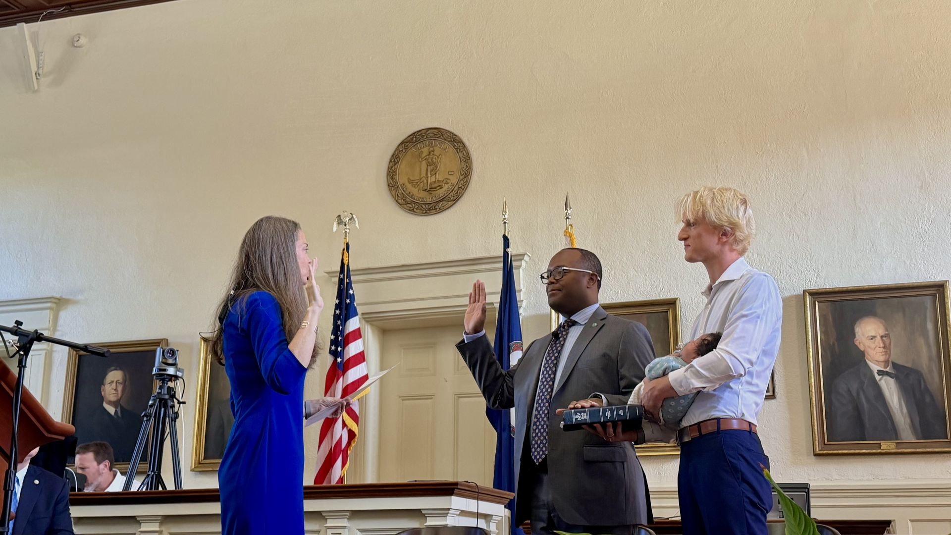 Oath ceremony in a formal room: a woman in blue and a man in a gray suit raise their hands; another man holds a baby. Flags, a Virginia seal, and portraits line the wall.