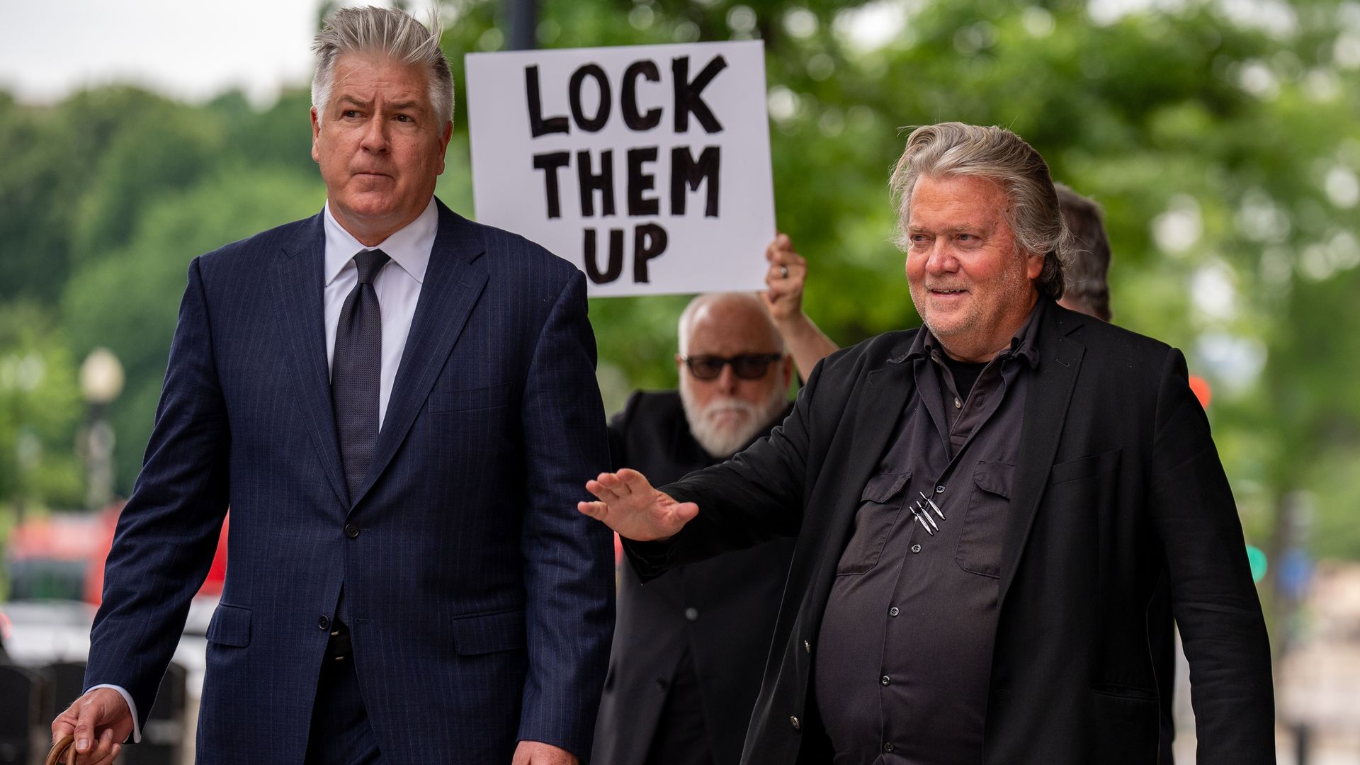 Steve Bannon, on the right and dressed in black, walks with his attorney as a protester behind them holds a sign that says "Lock Them Up."