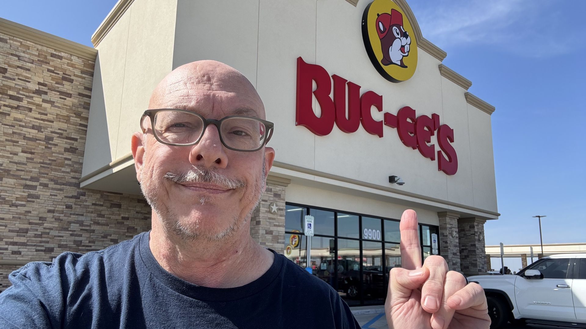 A photo of a bald man standing in front of a Buc-ee's store pointing at the sign.