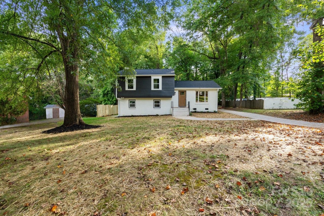 Two-story house with white lower walls and black upper siding surrounded by tall green trees, dry grass and fallen leaves on yard, sunny day with blue sky.