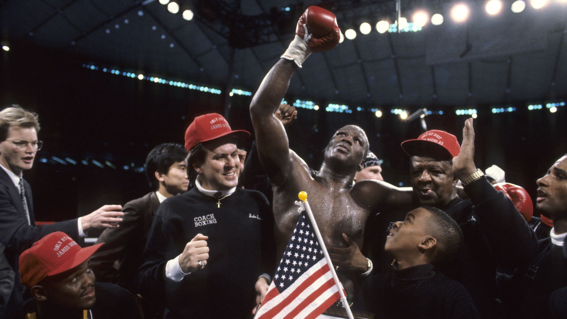 Boxer James "Buster" Douglas, in the center, raises a gloved fist in victory, surrounded by cheering supporters in an arena; an American flag and bright lights frame the scene.