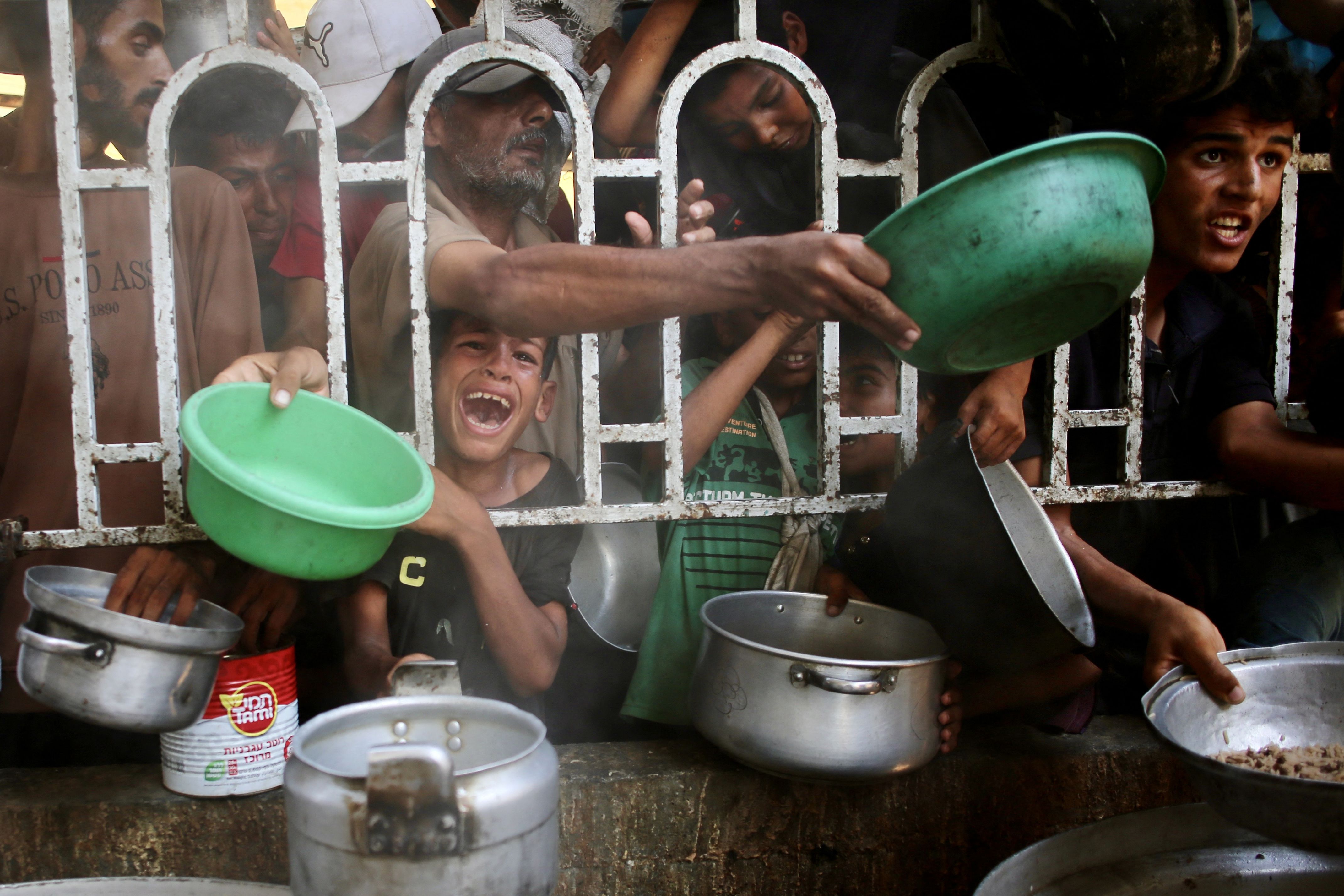 A group of men and boys behind metal bars eagerly hold out pots and bowls to receive food, showing expressions of urgency and need in a crowded setting.
