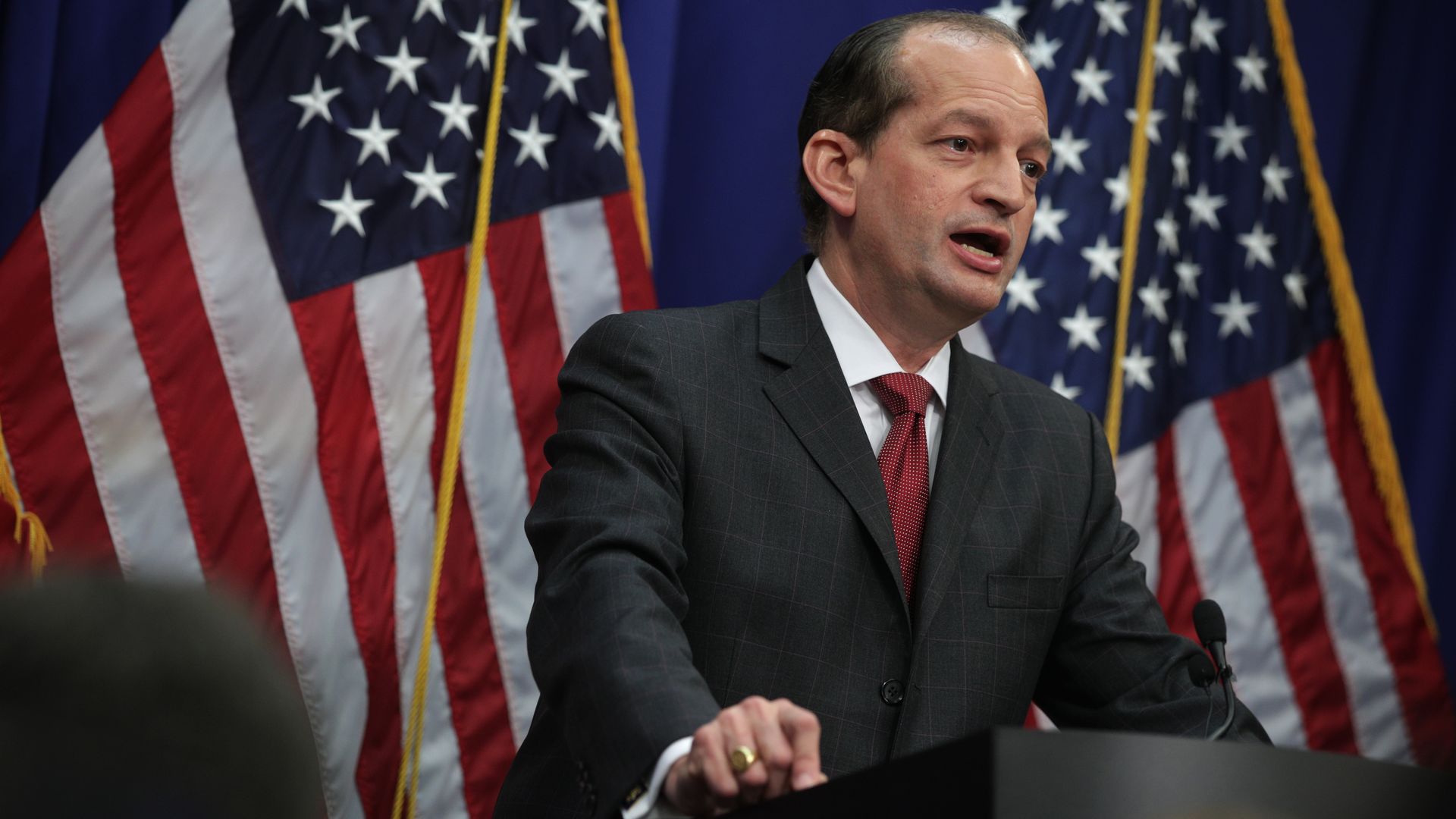 Photo of Alex Acosta standing at a podium with two American flags behind him
