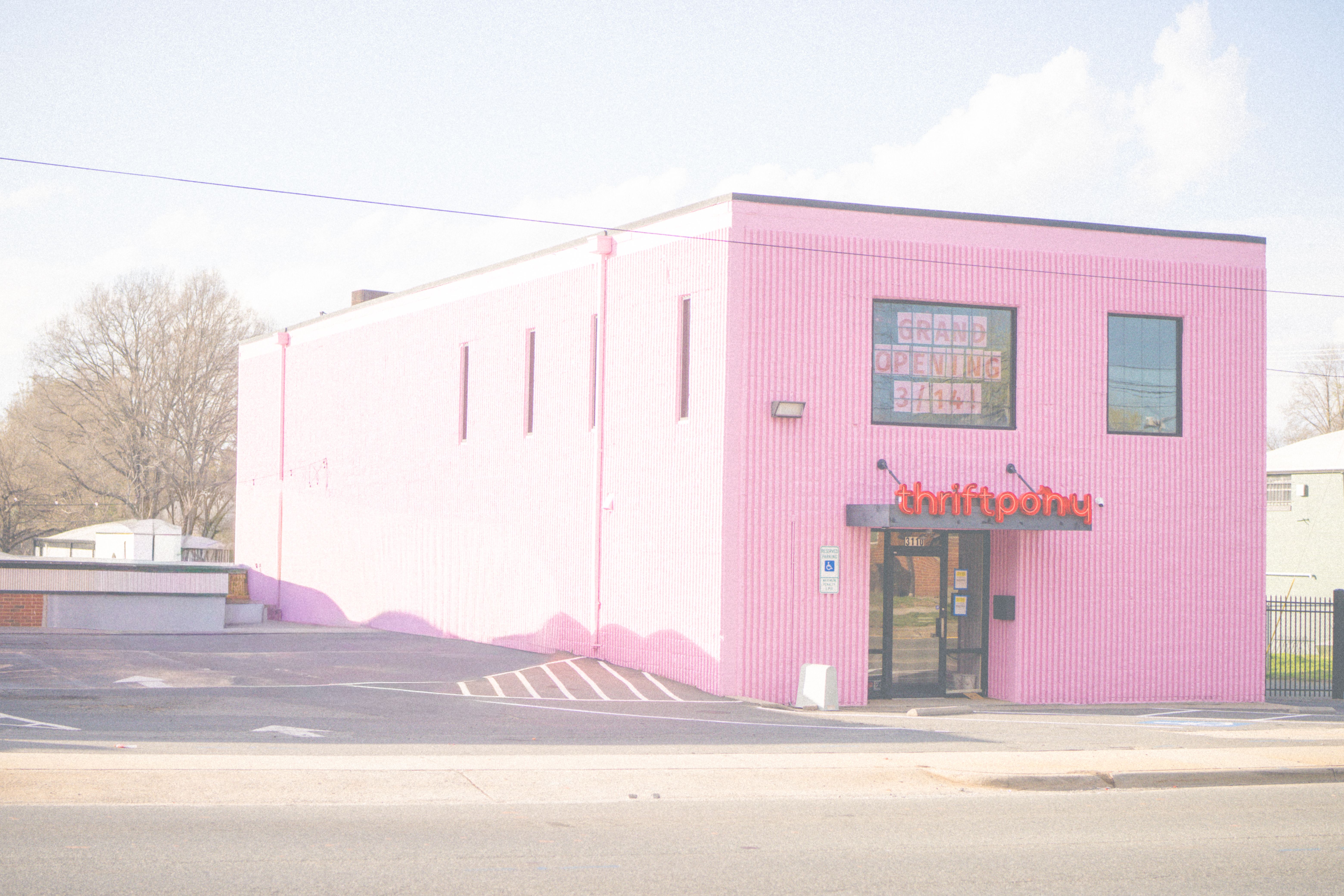 Pink corrugated storefront with a glass door and two windows; neon orange thriftpony sign above the entry. A GRAND OPENING window sign and a parking lot in front under a clear sky.