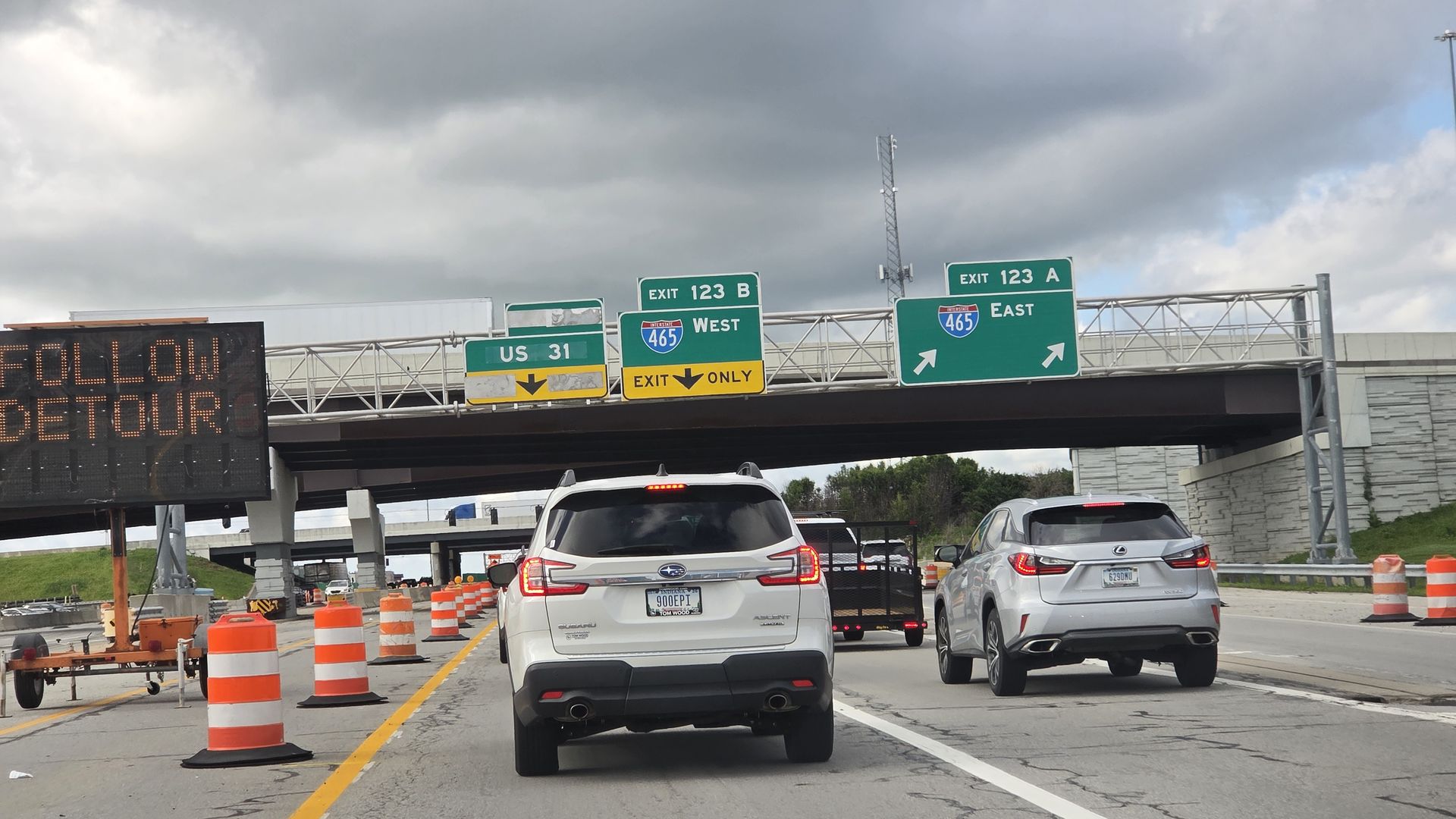 Cars on a highway beneath an overpass, with green signs for Exit 123A East, Exit 123B West, US 31, and I-465 East/West. Orange construction barrels line the road; an LED sign shows a detour message under a cloudy sky.