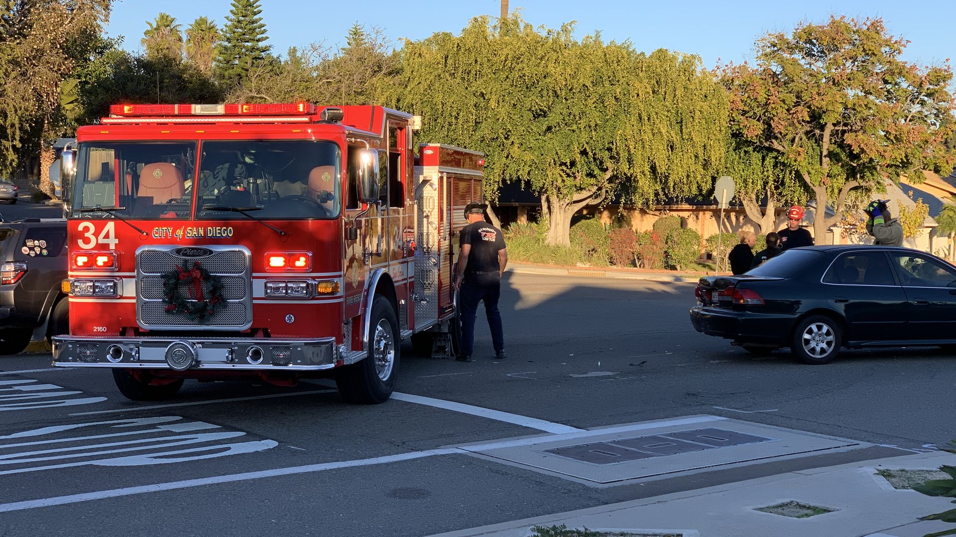 Red City of San Diego fire truck 34 with lights on at a street scene where firefighters and people stand beside a dark green car, sunny day with green trees in the background.