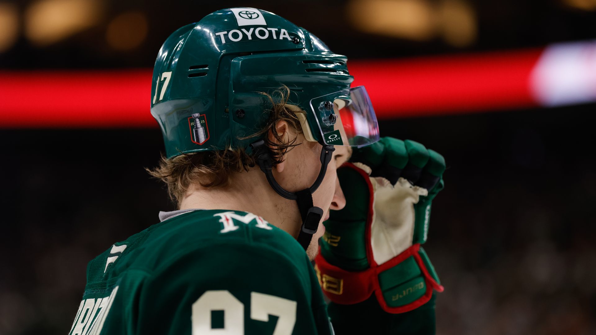 A hockey player in a green jersey with a 97 patch on the shoulder and a helmet raises his glove to adjust his helmet
