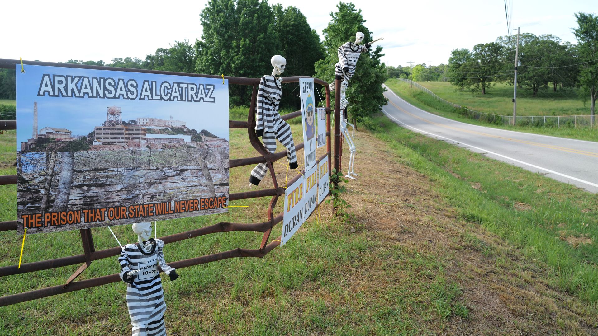Skeletons in prison stripes hang on a roadside fence with signs reading "Arkansas Alcatraz" and "Fire Duran Duran."
