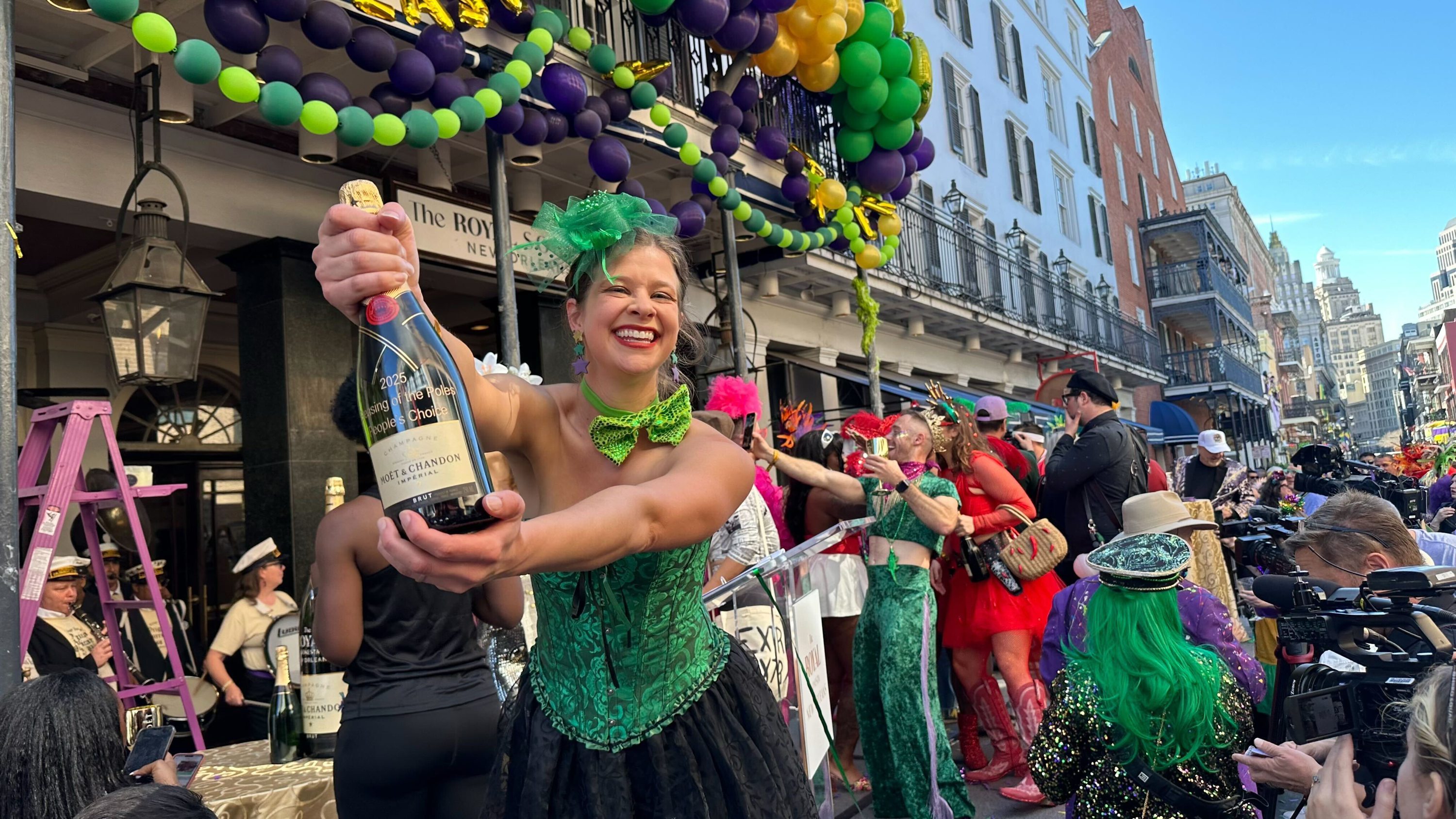 A woman wearing a green corset holds a bottle of champagne toward the camera and smiles.