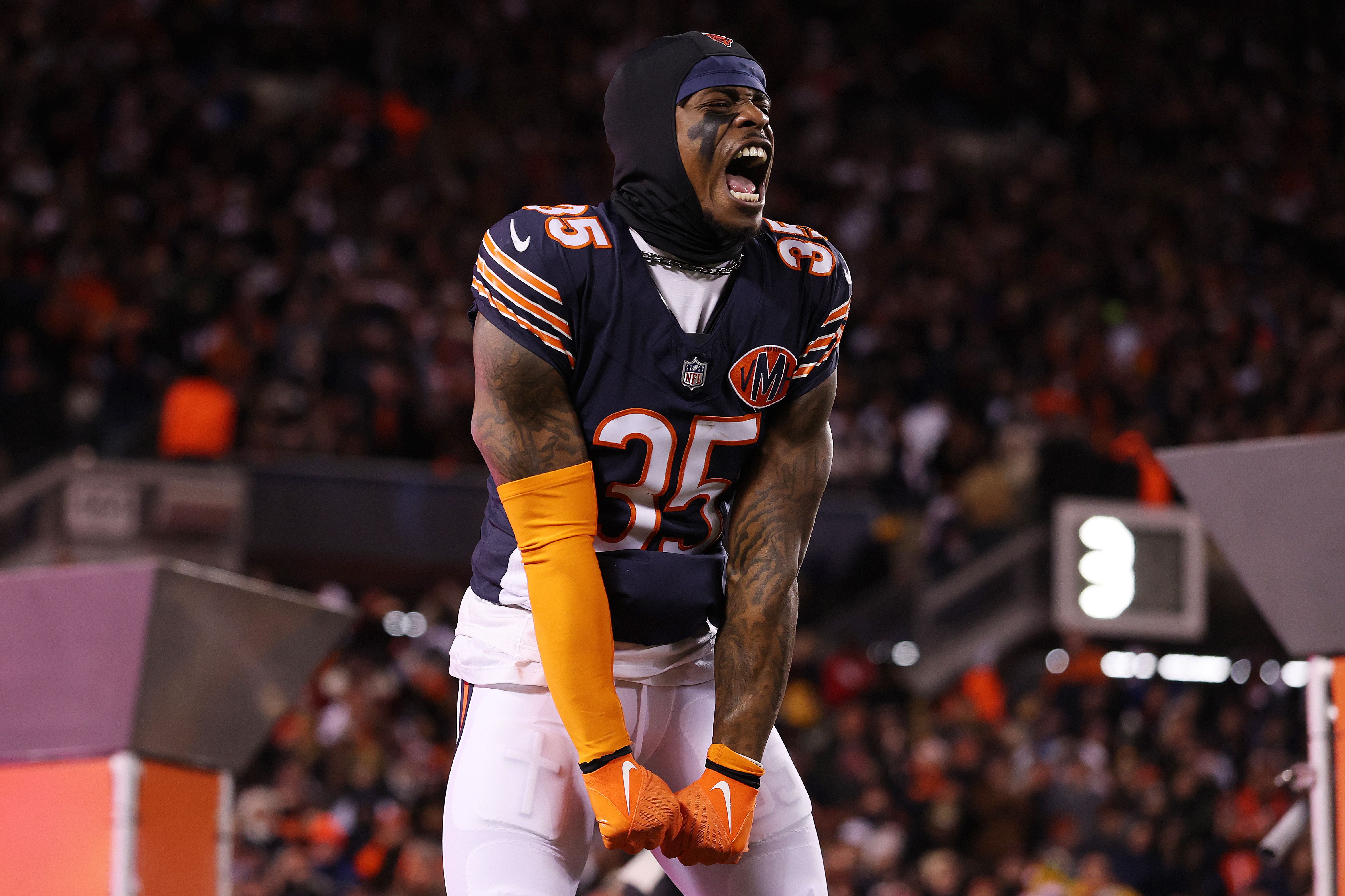 Chicago Bears football player in navy and orange uniform #35 shouting and clenching fists on field during game, with a crowd and scoreboard blurred in background.