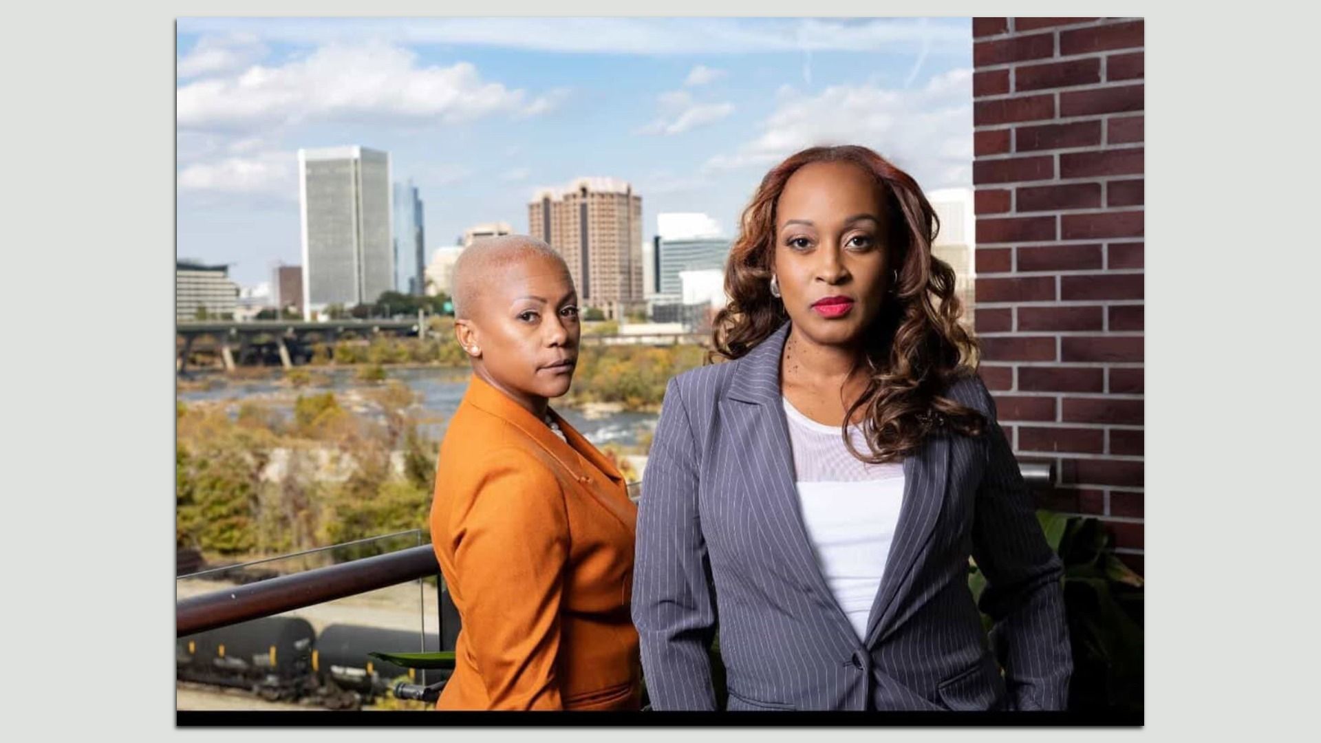 Two women, one in an orange blazer with short hair and the other in a gray pinstripe blazer with long curled hair, stand on a balcony overlooking a cityscape with tall buildings and a river.