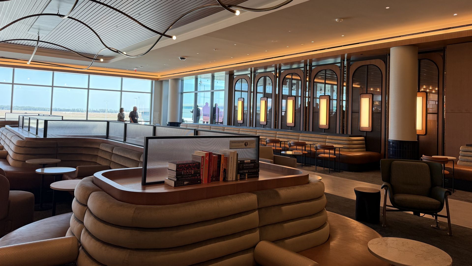 Airport lounge with circular beige sofa booths, a central table of books, and looping metal light fixtures overhead. Warm wall lighting and large windows overlook the tarmac.