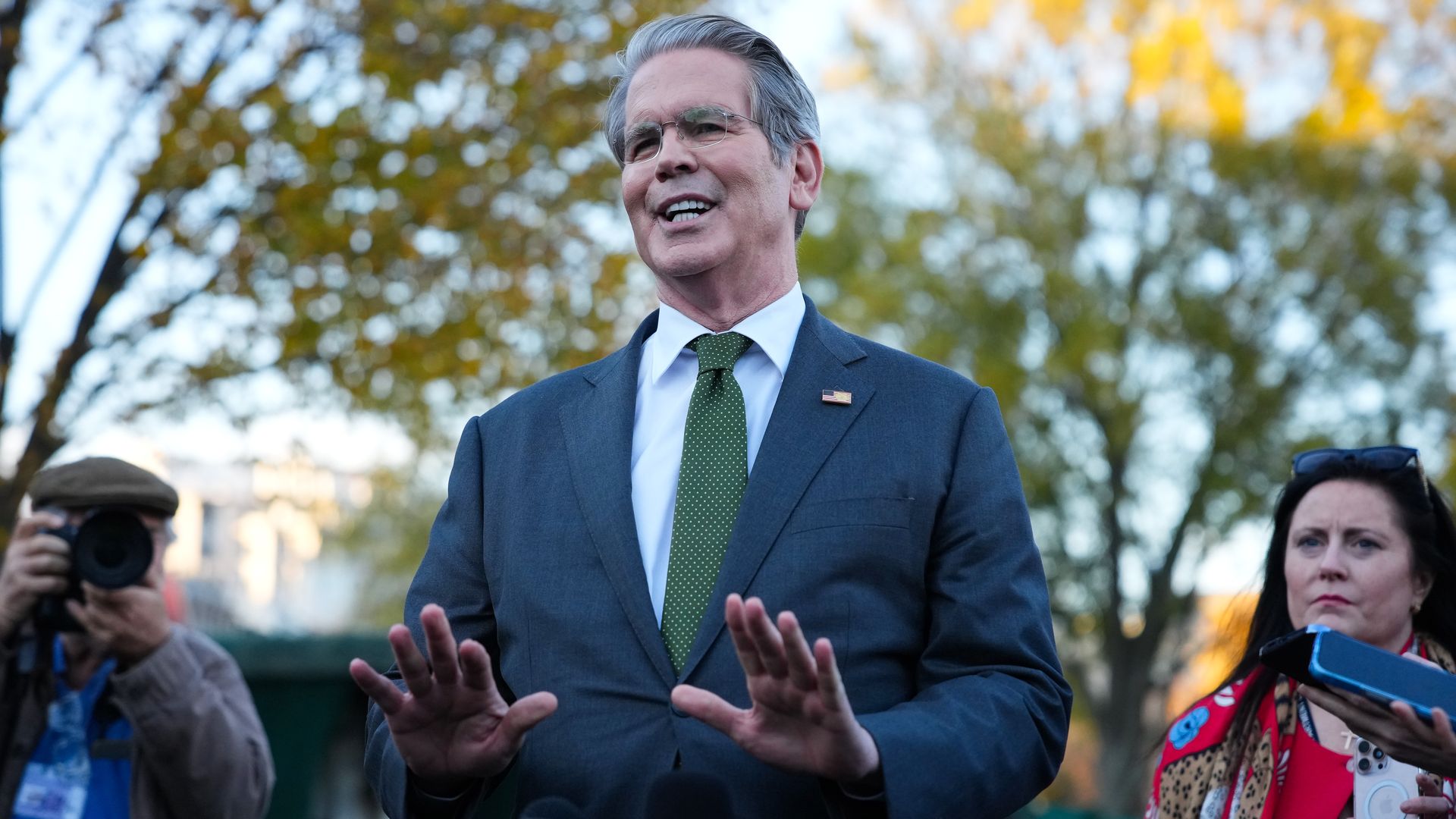 Treasury Secretary Scott Bessent, in a suit with green tie, speaks to reporters while making a downward-pushing gesture with both hands. 