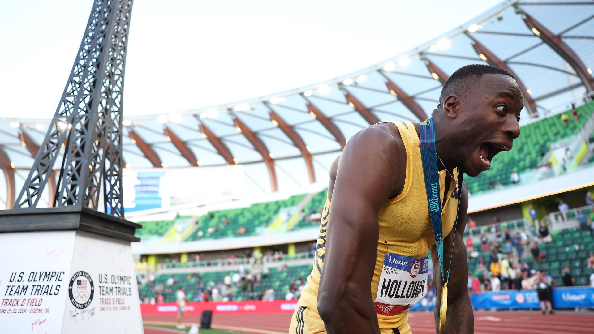 A smaller version of the eiffel tower on top of a thing that says "US olympic team trials" and a man excitedly yelling with a medal around his neck.