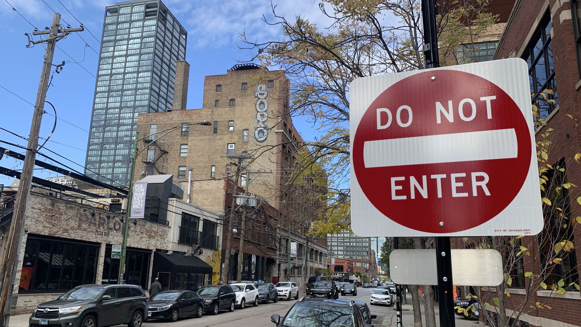 Red and white "DO NOT ENTER" traffic sign on a city street with parked cars, brick buildings, and a tall glass skyscraper under a blue sky with light clouds.