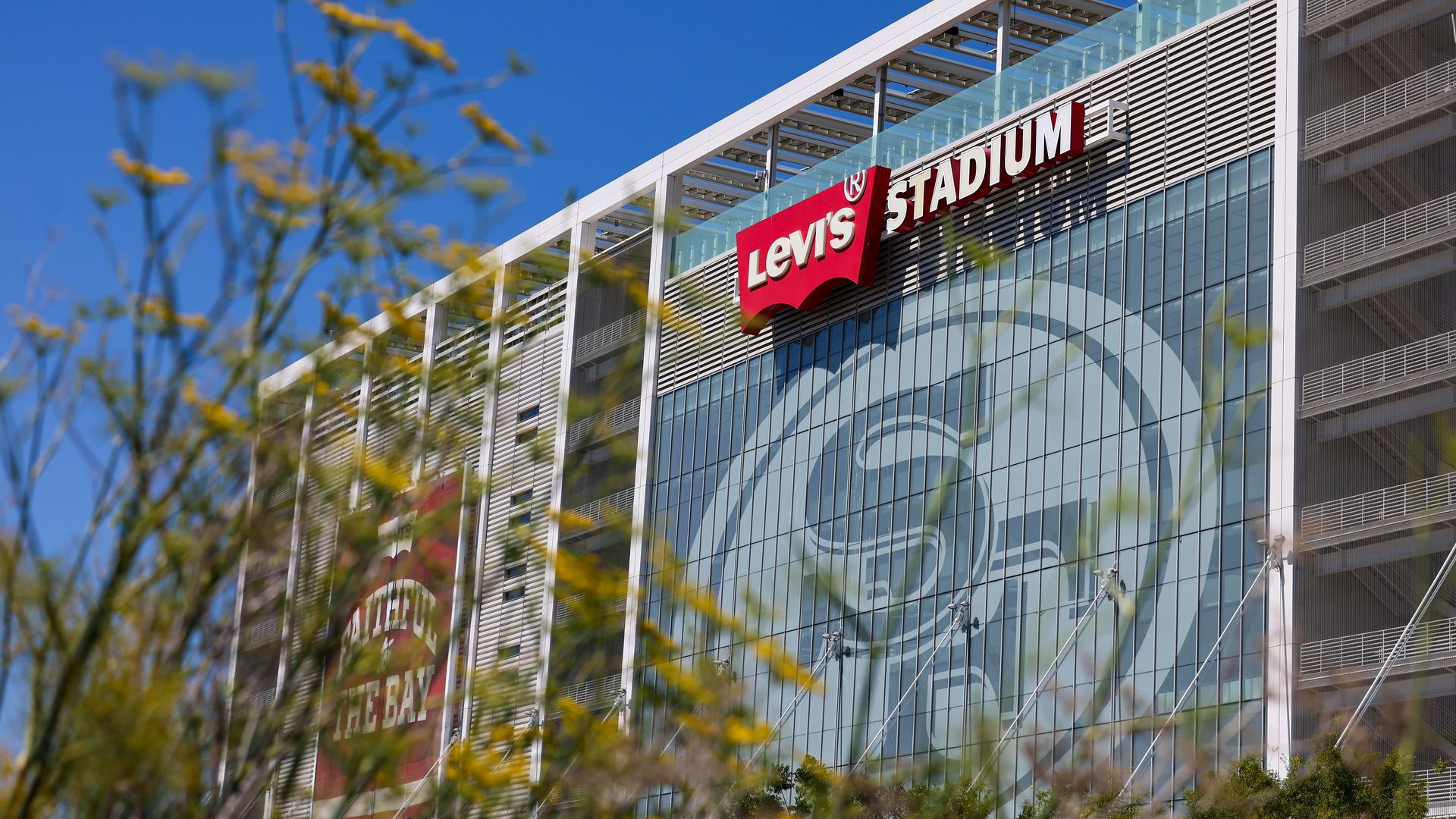 Levi's Stadium exterior with red logo sign, large 49ers emblem on glass facade, yellow wildflowers in foreground under clear blue sky.