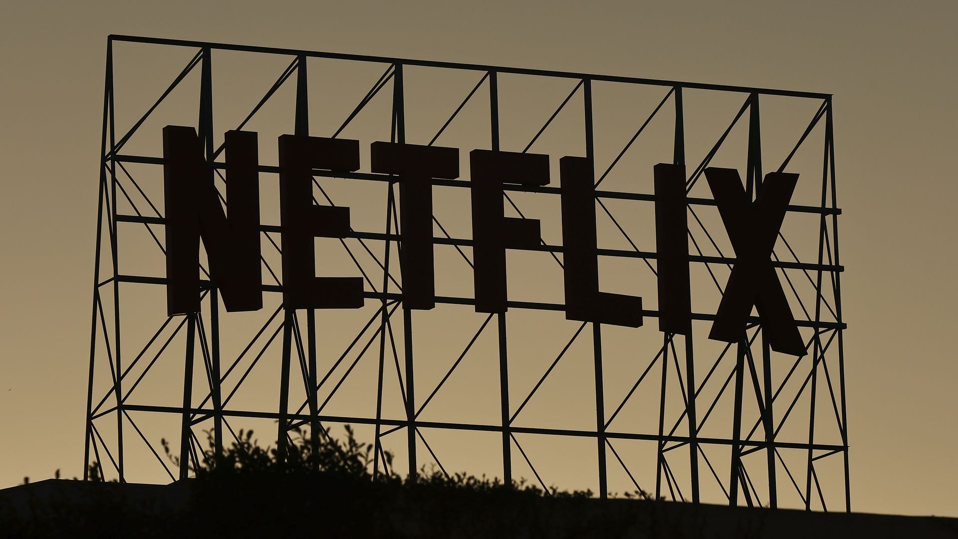 Silhouette of a large metal signframe with the word "NETFLIX" against a warm sunset sky; angular lattice supports and a dark hillside foreground.