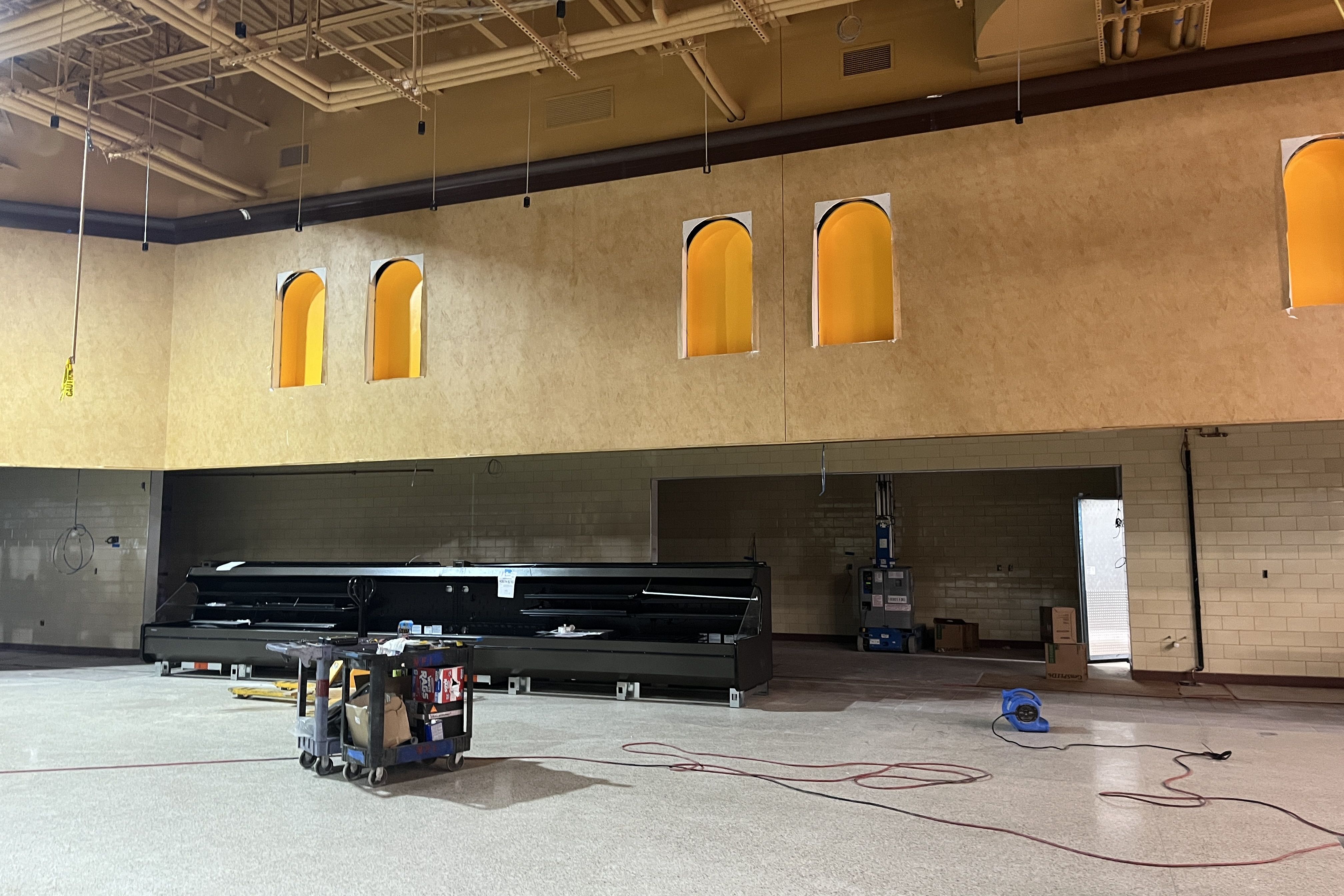Industrial interior under construction: beige wall with several bright yellow arched window openings, exposed ceiling pipes and wires, and carts with tools on a concrete floor.