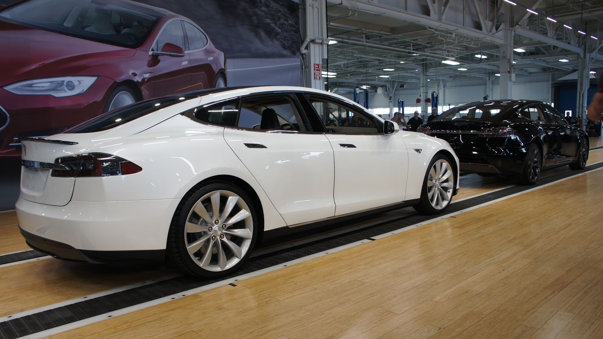 White and black Tesla Model S cars parked inside a spacious, brightly lit factory with wood flooring and people in the background.