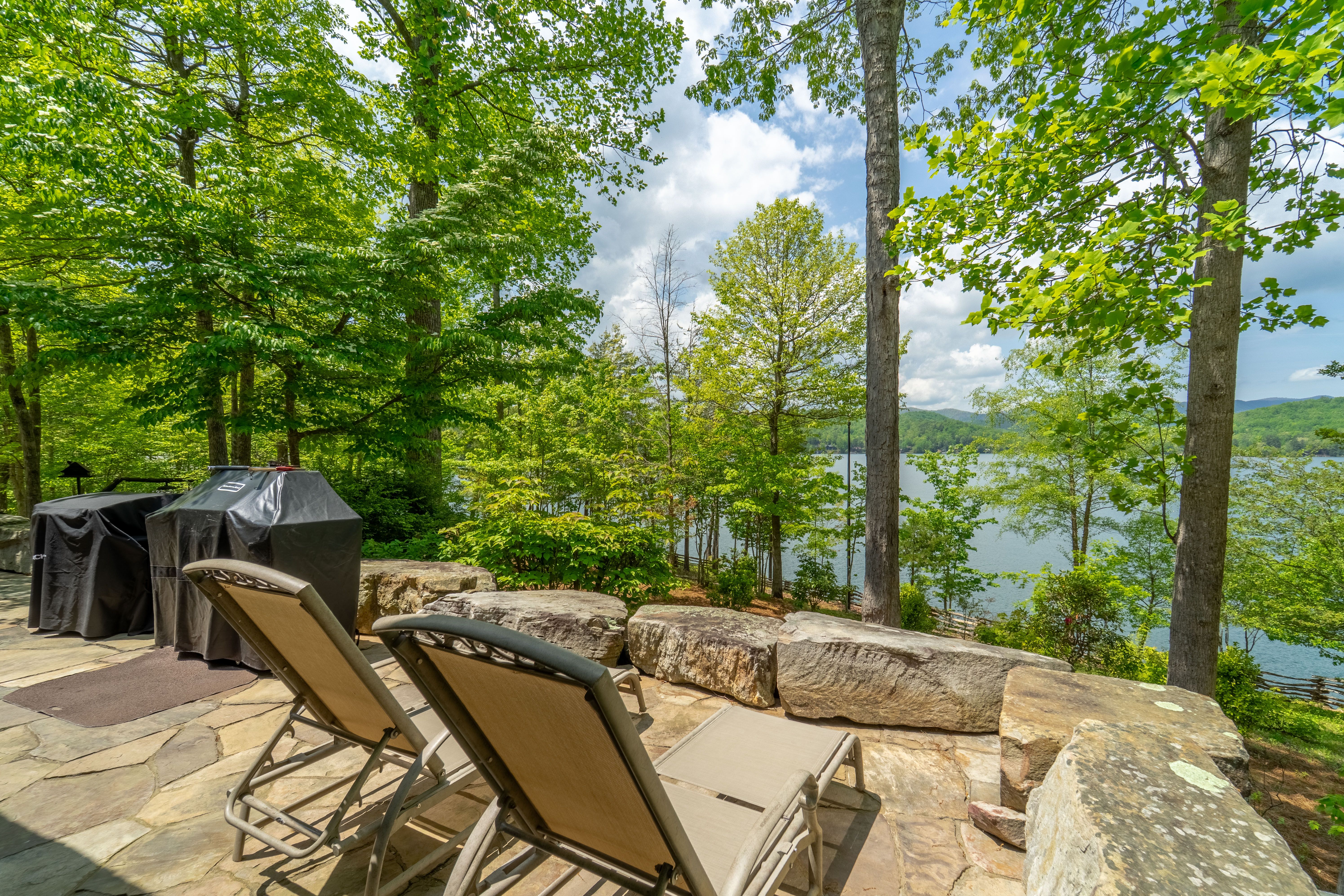 Two beige lounge chairs on stone patio surrounded by leafy green trees, overlooking a lake and distant mountains under a partly cloudy sky.